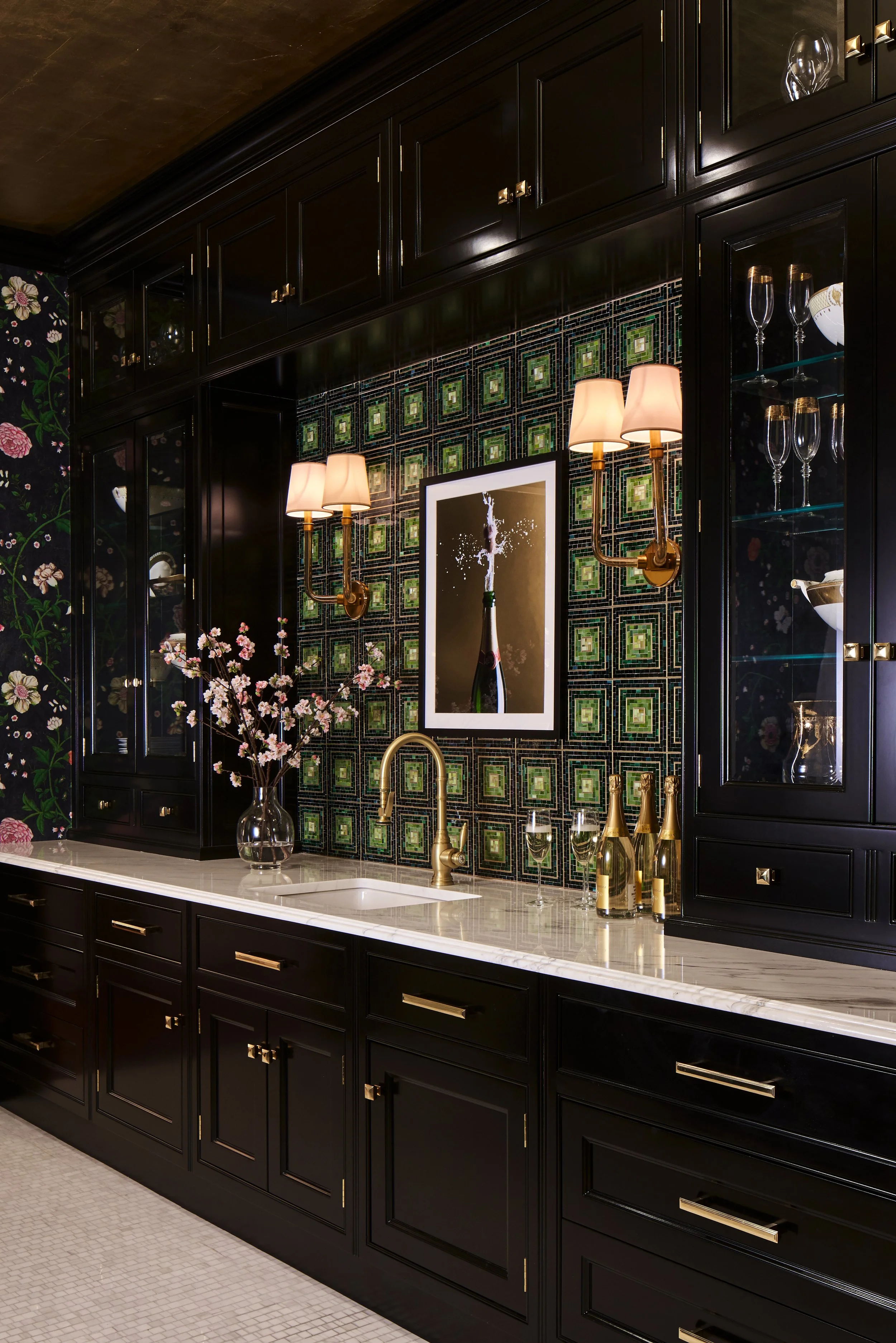 Black kitchen cabinets with gold hardware, a marble countertop, a bouquet of pink flowers, gold champagne bottles, a sink with a gold faucet, and a framed photo of a champagne bottle popping with splashes, on a green tiled backsplash with gold and bl