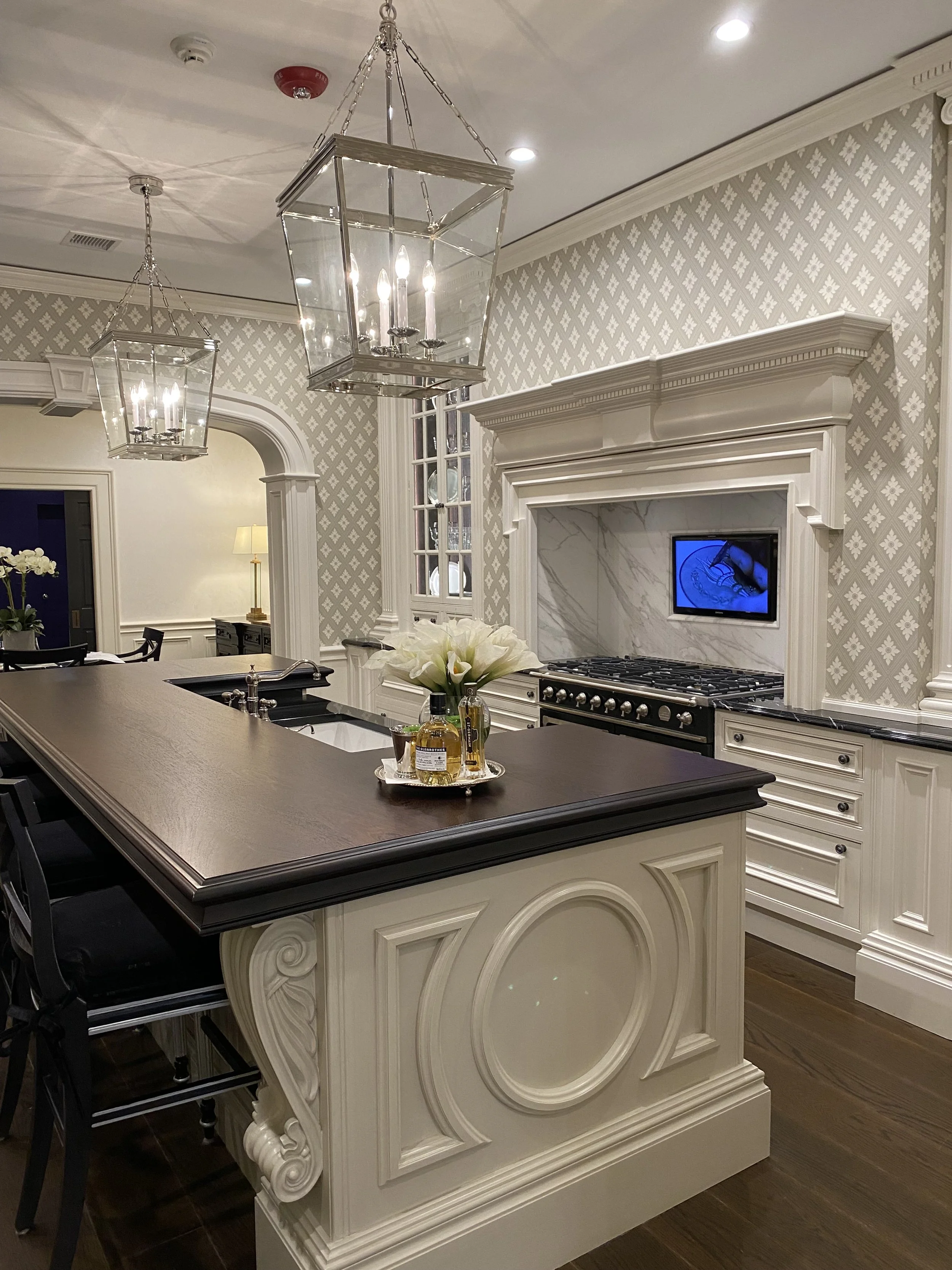 Elegant kitchen with a dark wood island, white cabinetry, marble backsplash, wall-mounted TV, floral centerpiece, and chandeliers.