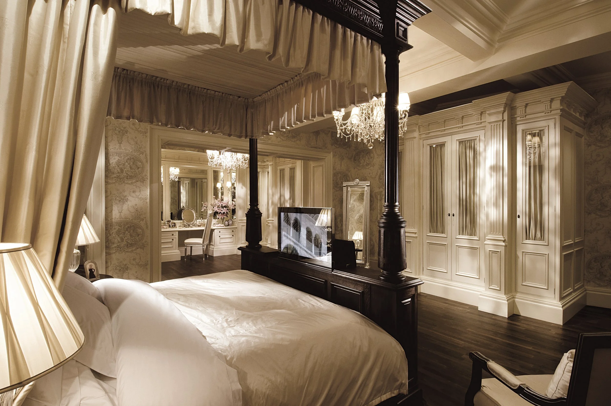 Luxurious bedroom with a four-poster canopy bed, elegant drapes, a chandelier, and a view into a dressing area with vanity table and mirror.