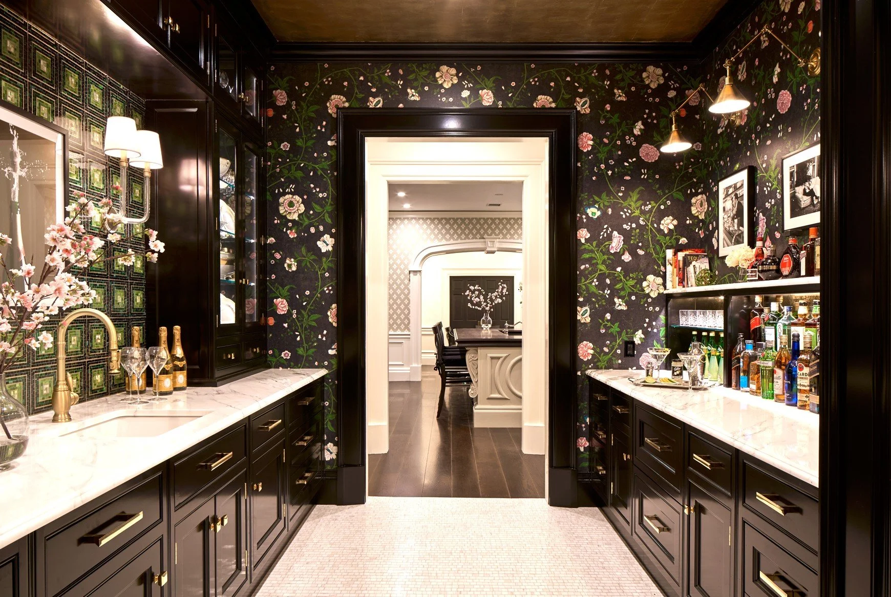 A luxurious bar area with black cabinetry, marble countertops, floral wallpaper, and bottles of liquor, leading to a dining room with dark wood furniture and floral arrangements.