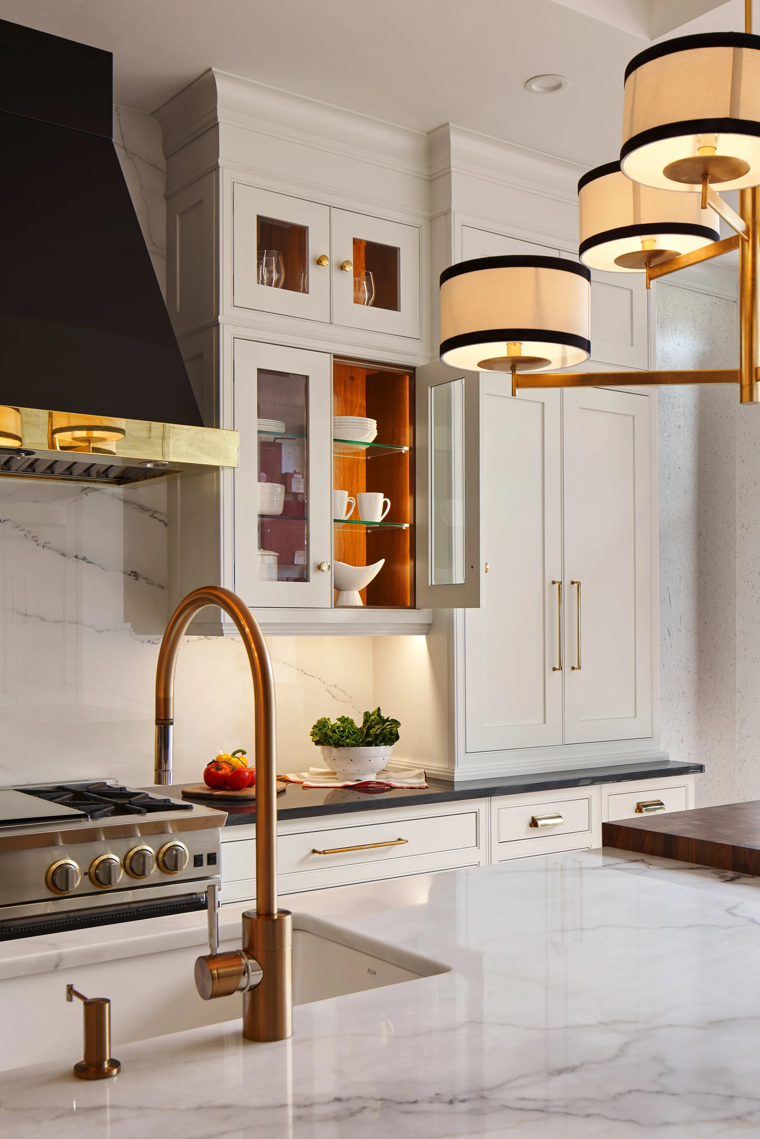 Modern kitchen with white cabinetry, marble countertops, brass accents, a copper faucet, a gas stove, open shelves with dishes, and a decorative light fixture.