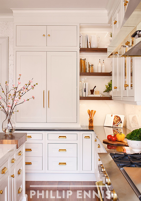 White kitchen cabinets with gold handles, open shelves with books and kitchen accessories, a bowl of vegetables, and a stove with a wooden cutting board.