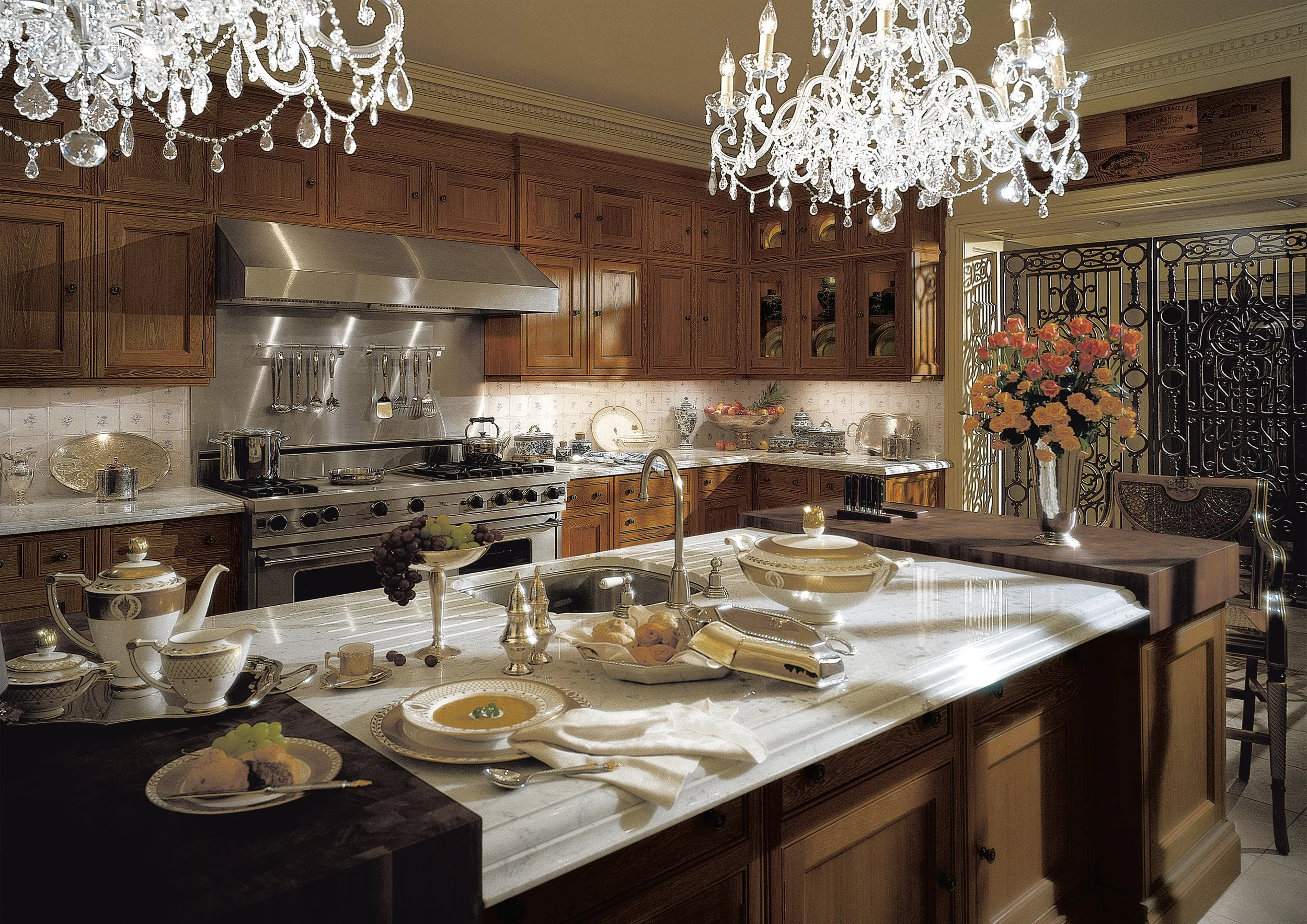 A luxurious kitchen with wooden cabinets, a marble island, a stainless steel stove, elegant chandeliers, and a large floral arrangement.