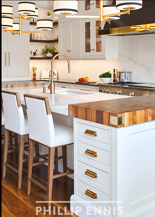 Modern kitchen with white cabinets, a marble island, and a wooden cutting board. Oversized black, white, and gold pendant lights hanging above the island. The kitchen has a stainless steel stove, a nickel faucet, and fresh vegetables on the counter.
