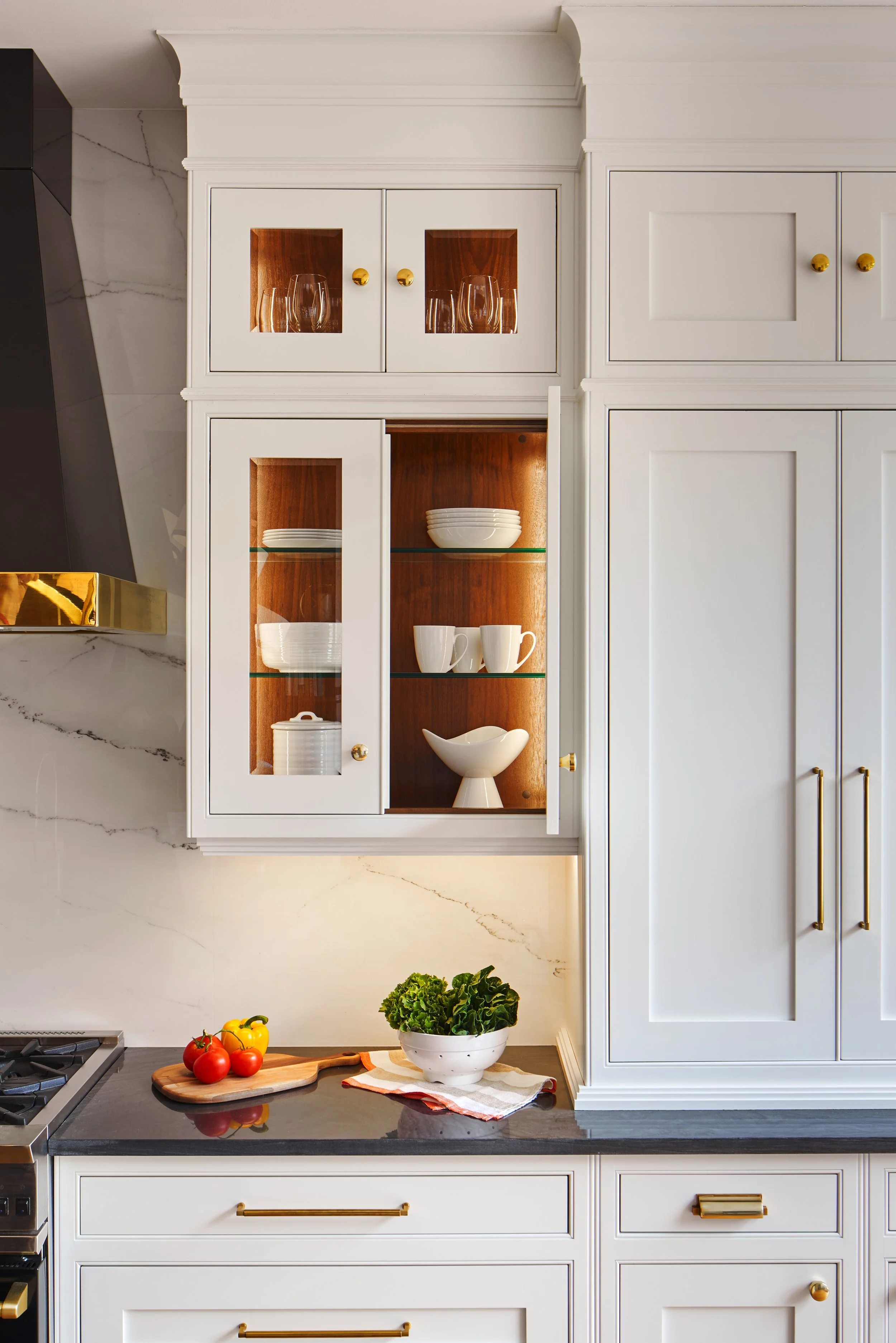White kitchen cabinet with glass doors displaying dishes and glassware, countertop with tomatoes, bell pepper, and a bowl of greens, marble backsplash, part of a stove on the left.
