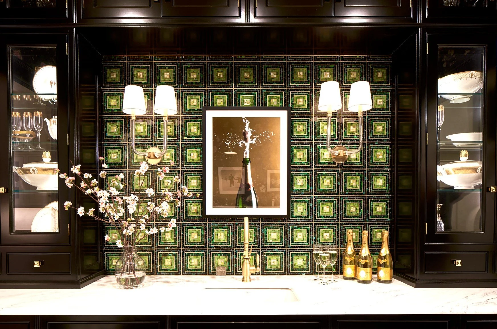 A kitchen or bar area with a black cabinet and glass display cases on either side. In the center, there is a white countertop with a vase holding flowering branches, a gold candle holder with a lit candle, two champagne bottles, and two glasses of ch