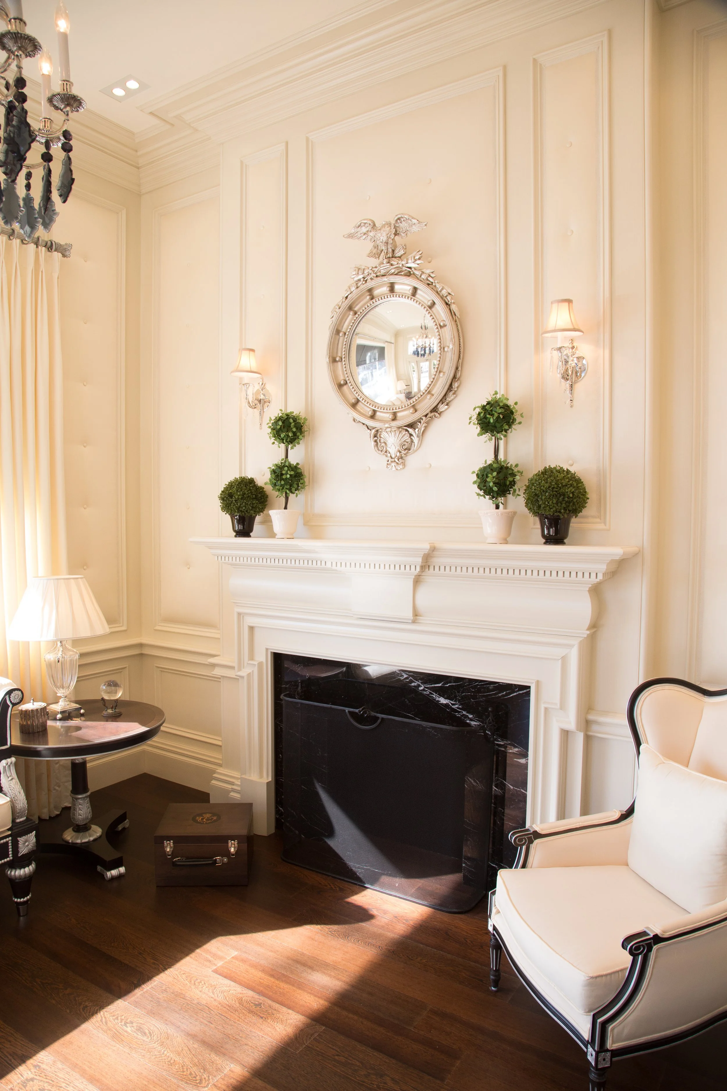 Elegant living room with cream walls, ornate fireplace, round mirror, wall sconce lights, small potted topiary plants, a white armchair, and a side table with a lamp and glass decor, hardwood flooring.