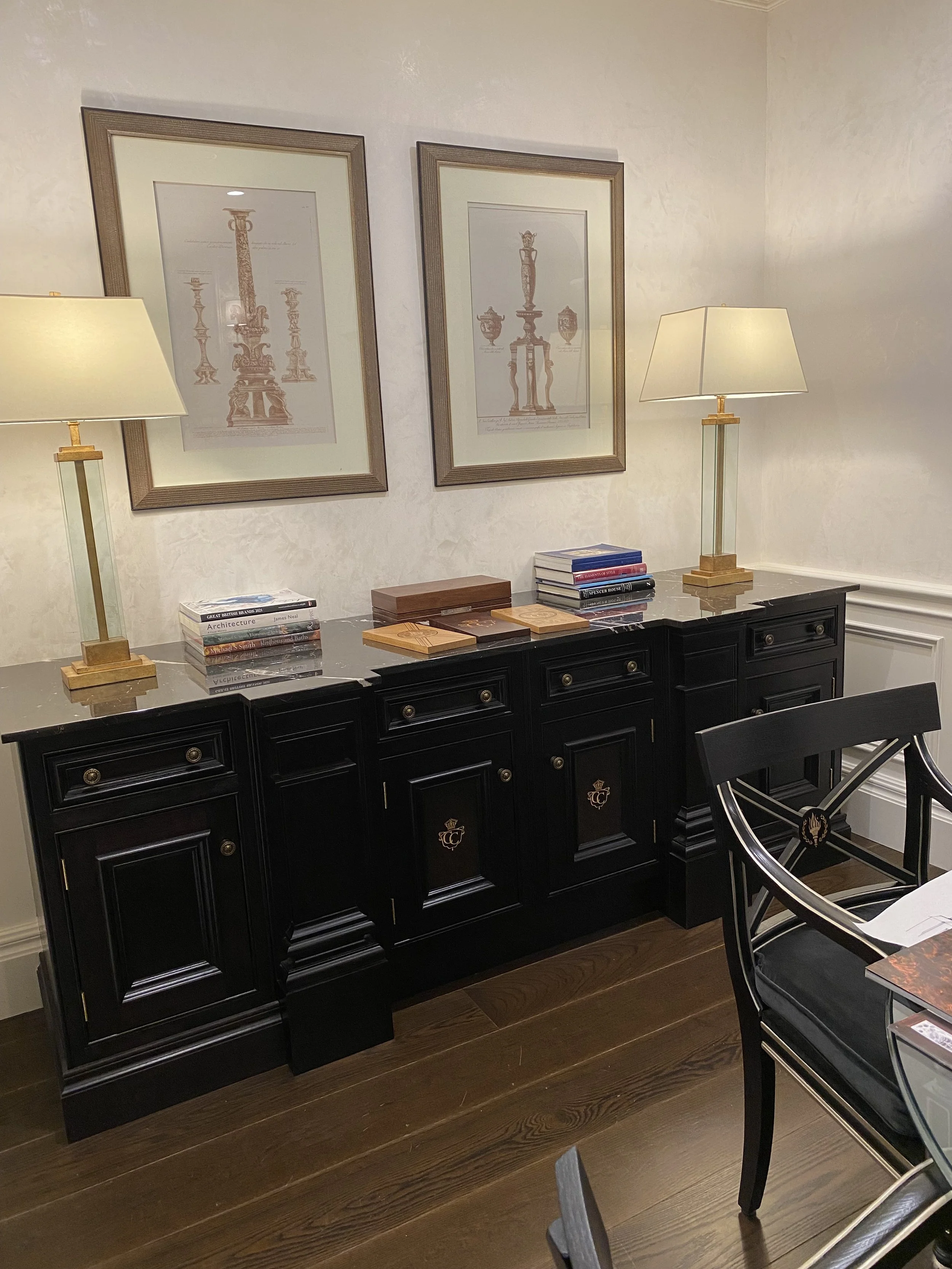 A black sideboard with gold accents and two white lamps on top, placed in a room with wood flooring. Above the sideboard are two framed prints of classical urns or vases, and on the sideboard are stacked books and wooden decorative items.