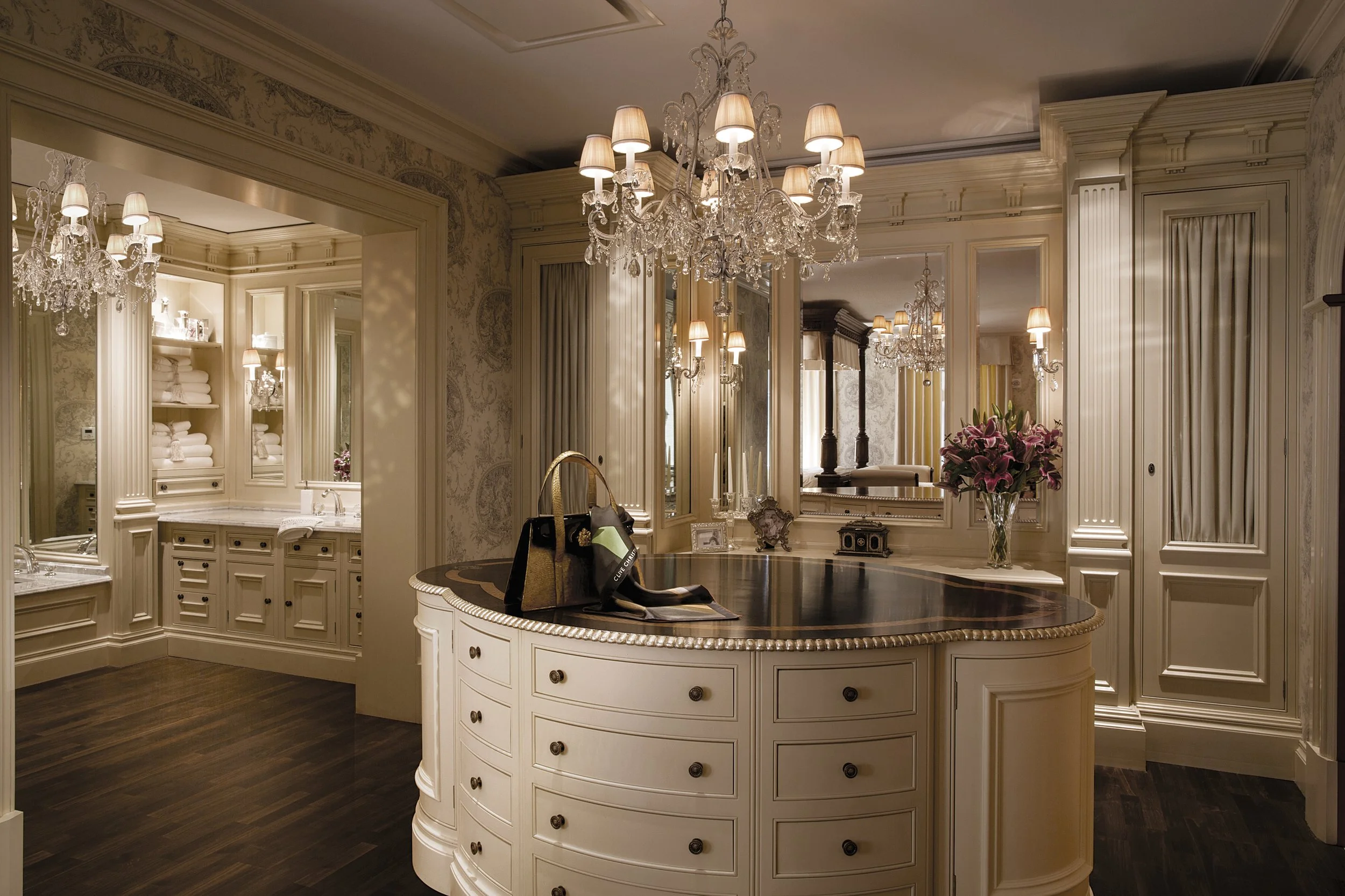 Luxury bathroom with ornate white cabinetry, large mirrors, chandelier lighting, and a dark marble-topped island with a leather handbag and wallet on it, decorated with pink lilies in a vase.