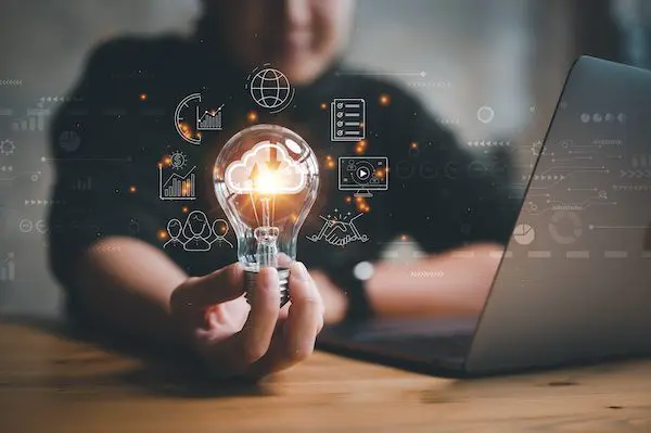 Person holding a light bulb with cloud and digital technology icons around it, sitting at a desk with a laptop.
