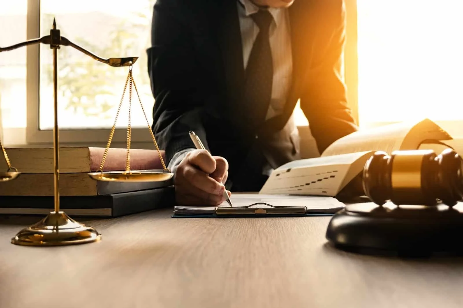 A person in a suit writing on a clipboard at a desk with legal books, a gavel, scale of justice, and documents, illuminated by natural light from a window.