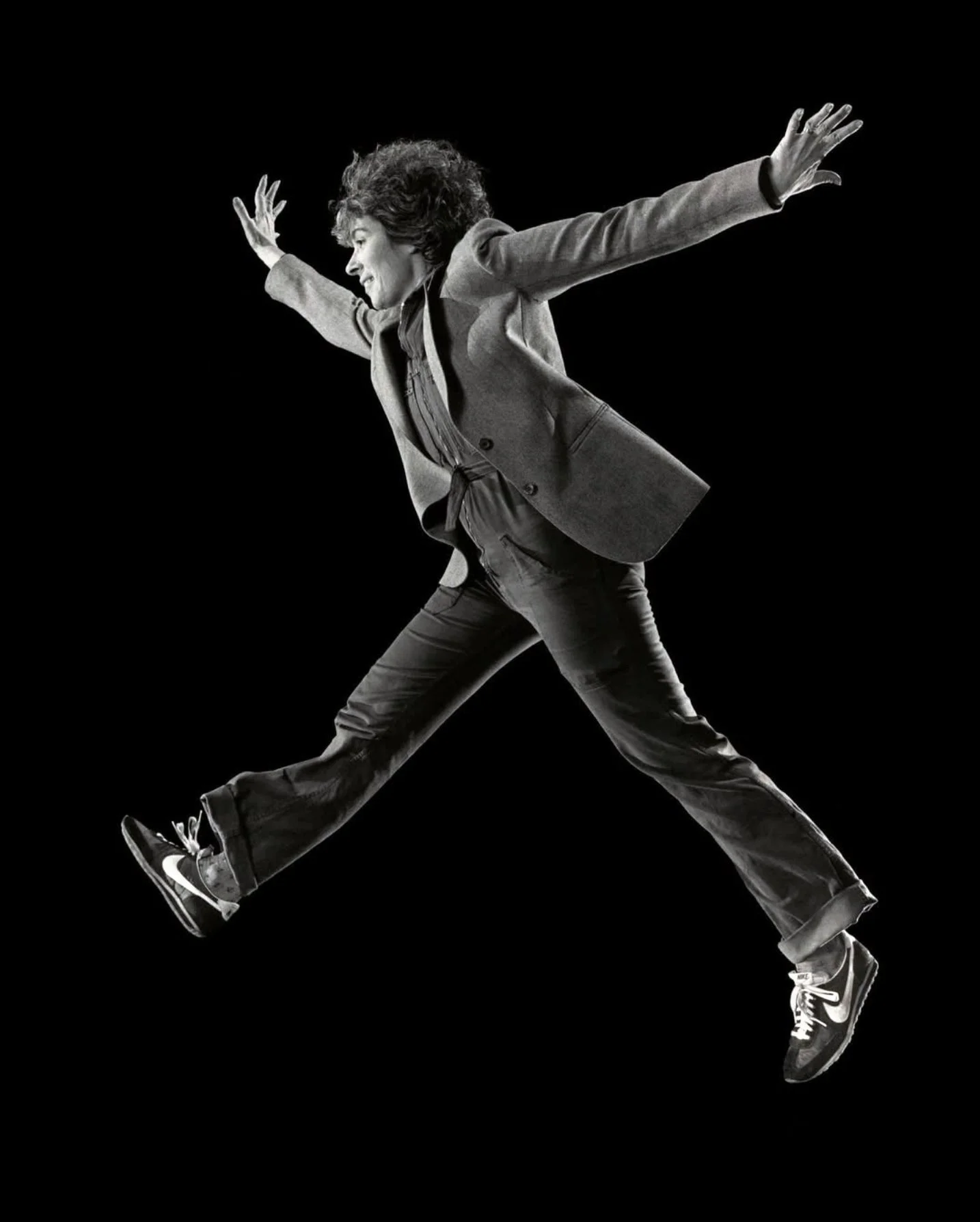 a middle aged woman in jeans and a blazer jumps high in the air against a dark background with arms and legs akimbo