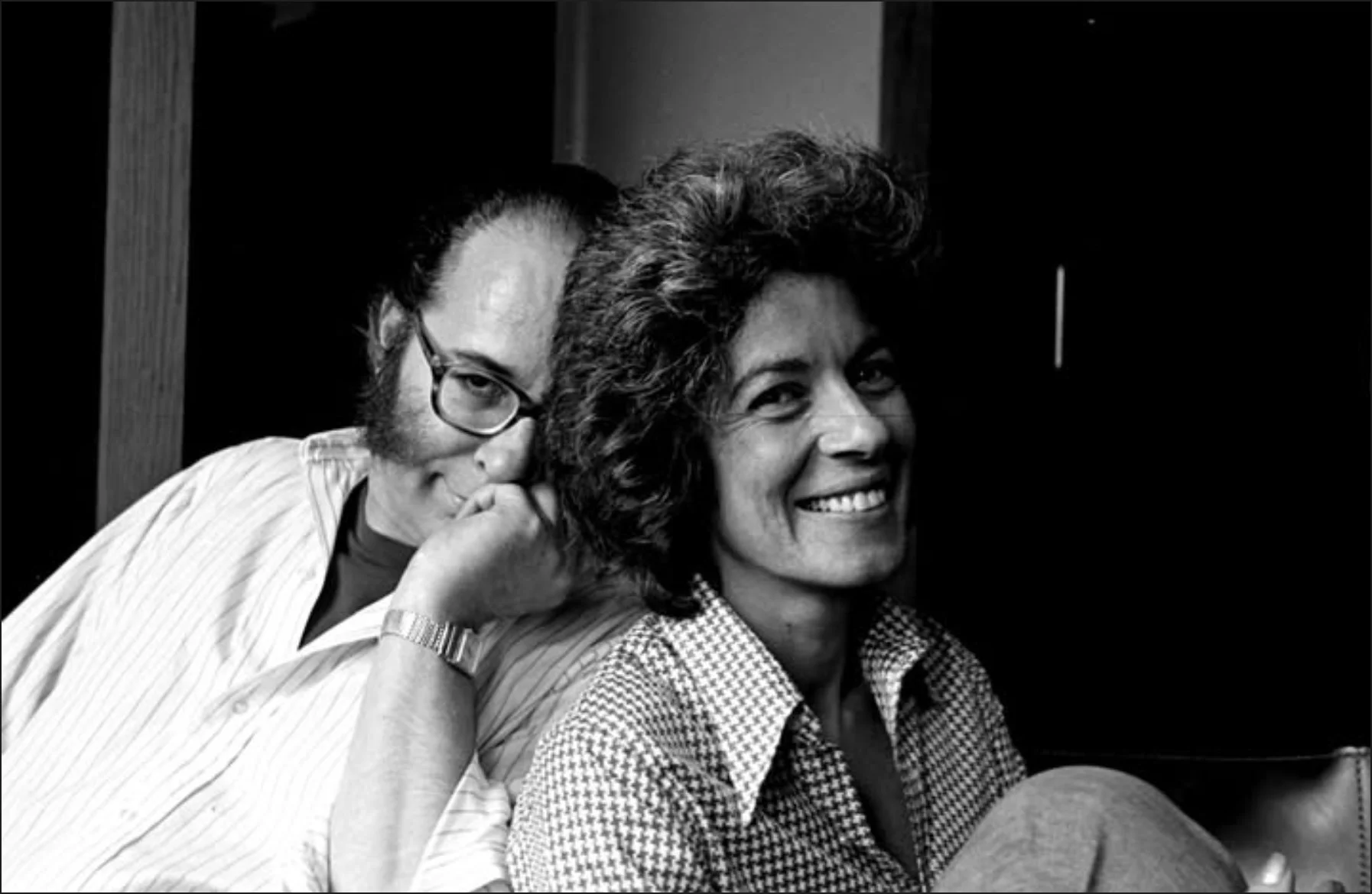 black and white photo of a middle aged couple, with the woman seated casually and smiling and the man also seated and grinning while leaning on her back