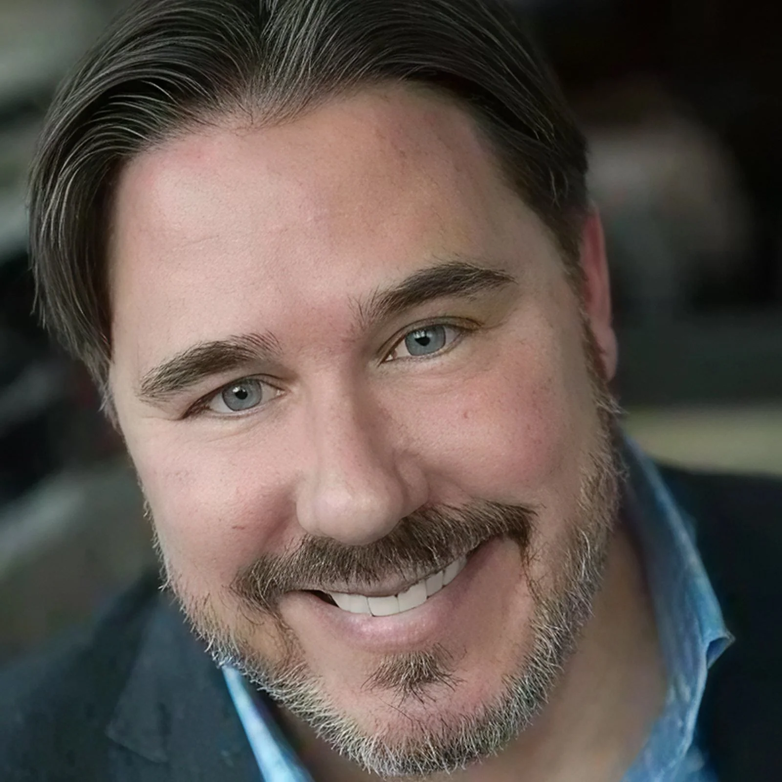 Close-up of a smiling man with blue eyes, brown hair, and a beard, wearing a dark blazer and light blue shirt.