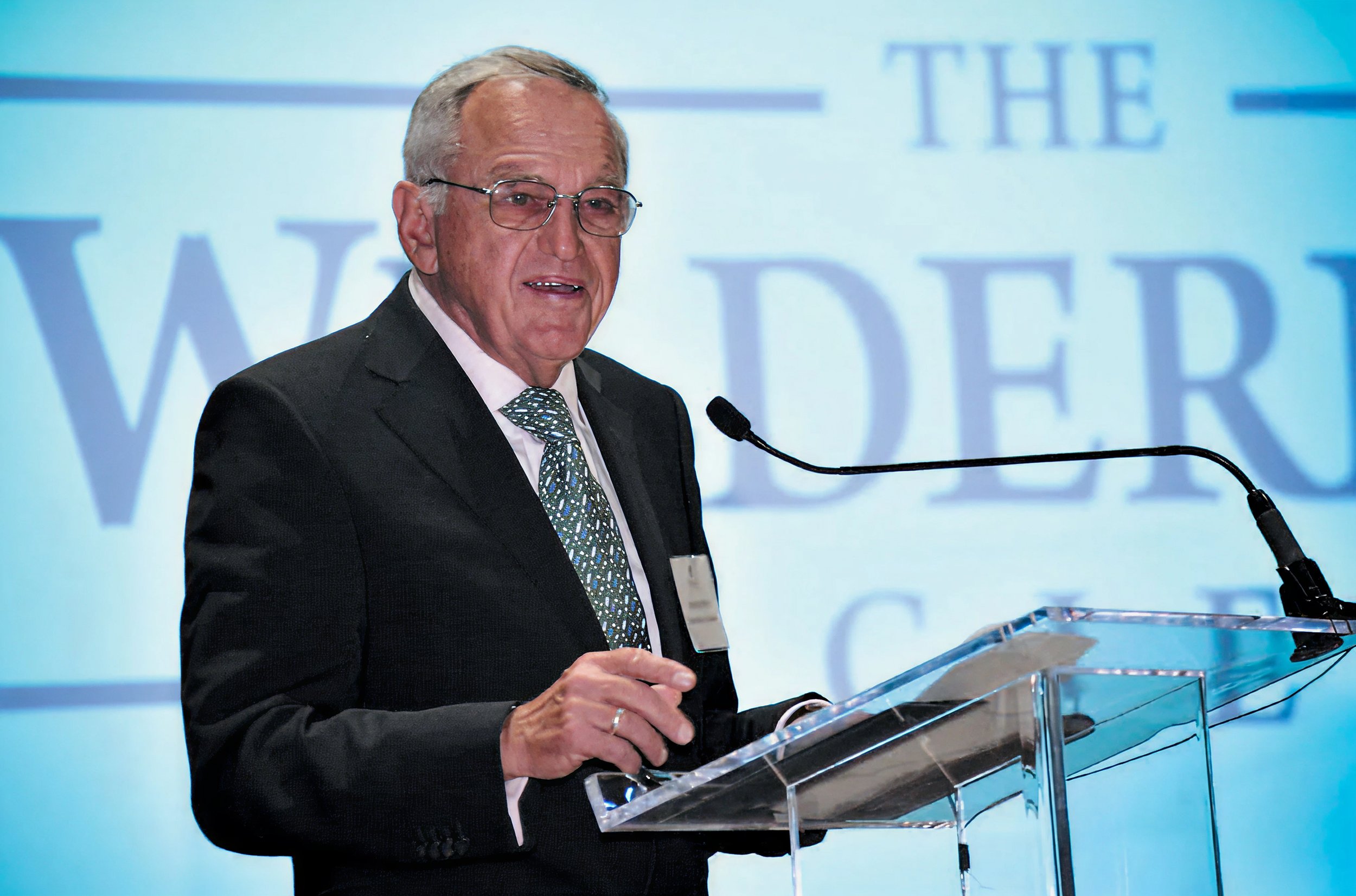 An older man with gray hair, glasses, wearing a black suit, white shirt, and a patterned tie, speaking at a podium with a microphone in front of a light blue background with large text.