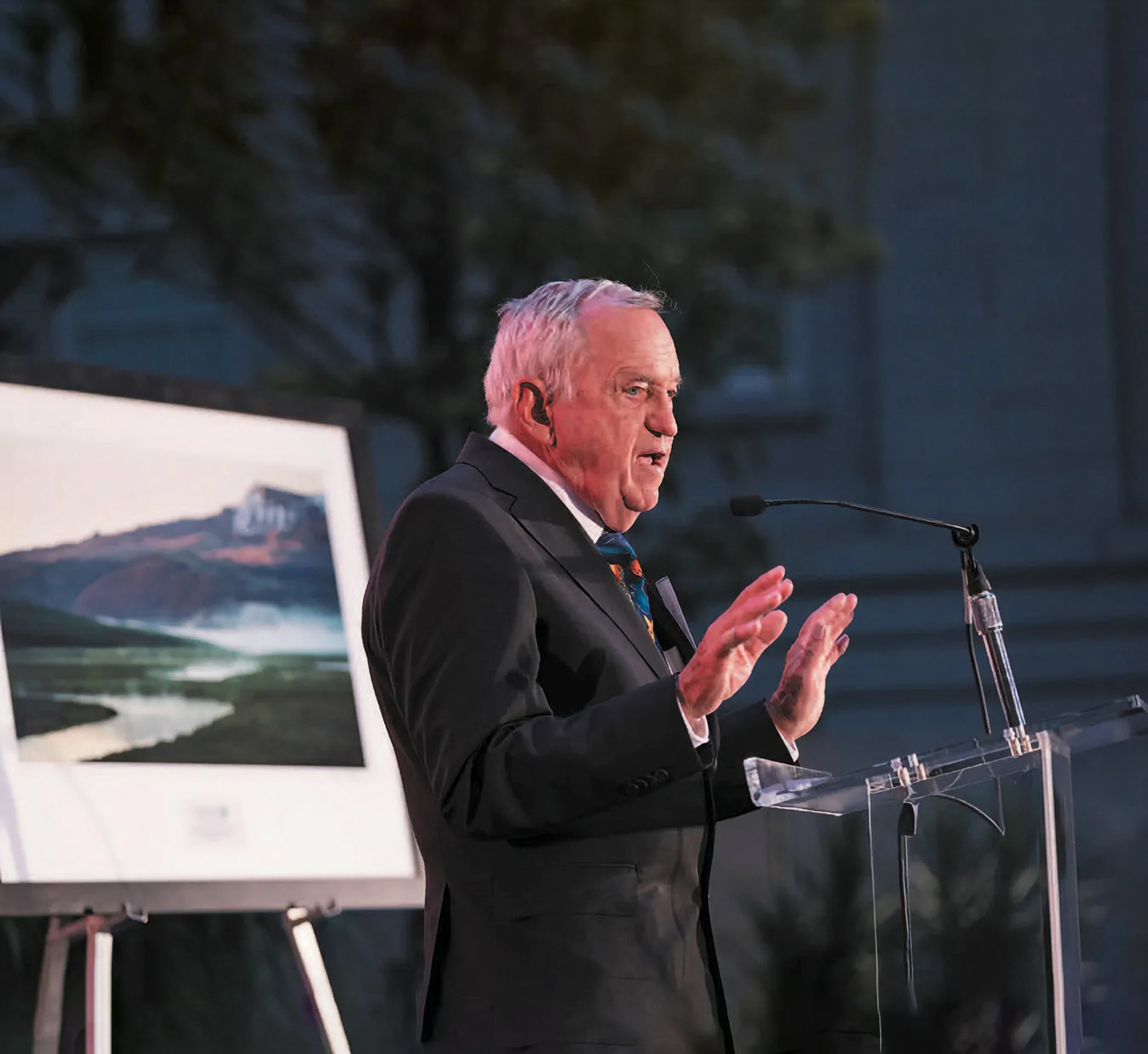 An older man in a black suit and tie is speaking at a clear acrylic podium with a microphone, gesturing with his hands. Behind him, there is a framed landscape painting of a river, mountains, and a distant building, on an easel, with a dark outdoor background.