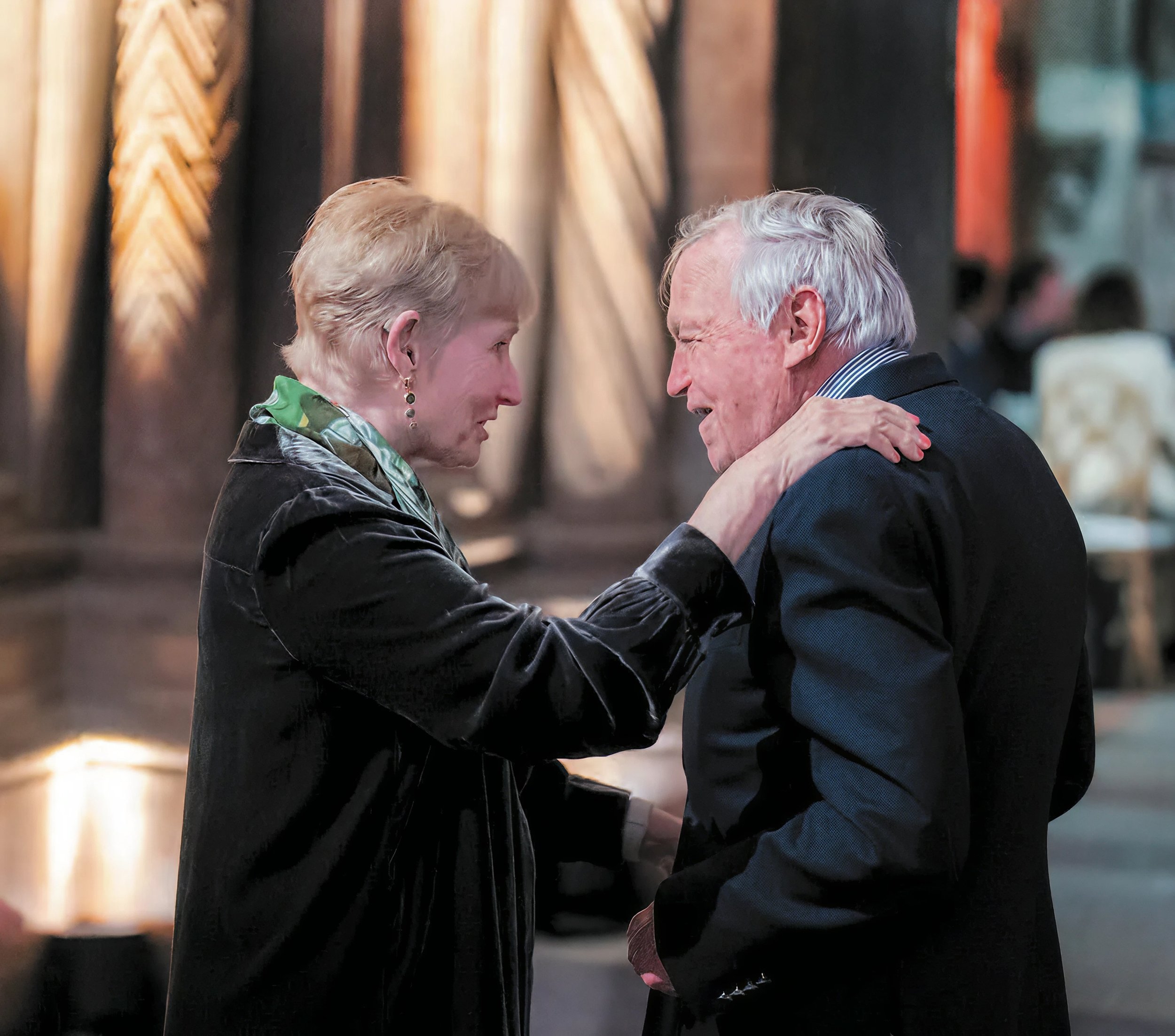 An elderly man and a woman are sharing a warm moment, smiling and looking into each other's eyes, with the woman touching the man's shoulder.