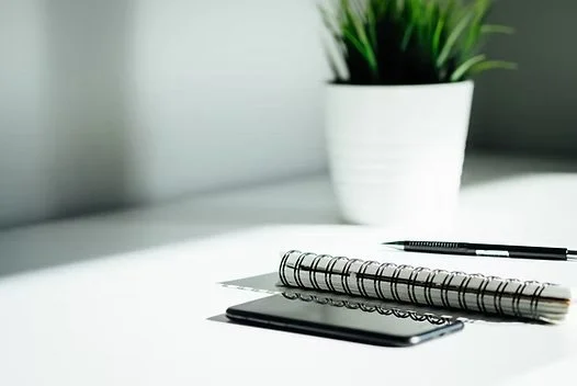 A white desk with a potted green plant, a black smartphone, a spiral notebook, and a black pen.