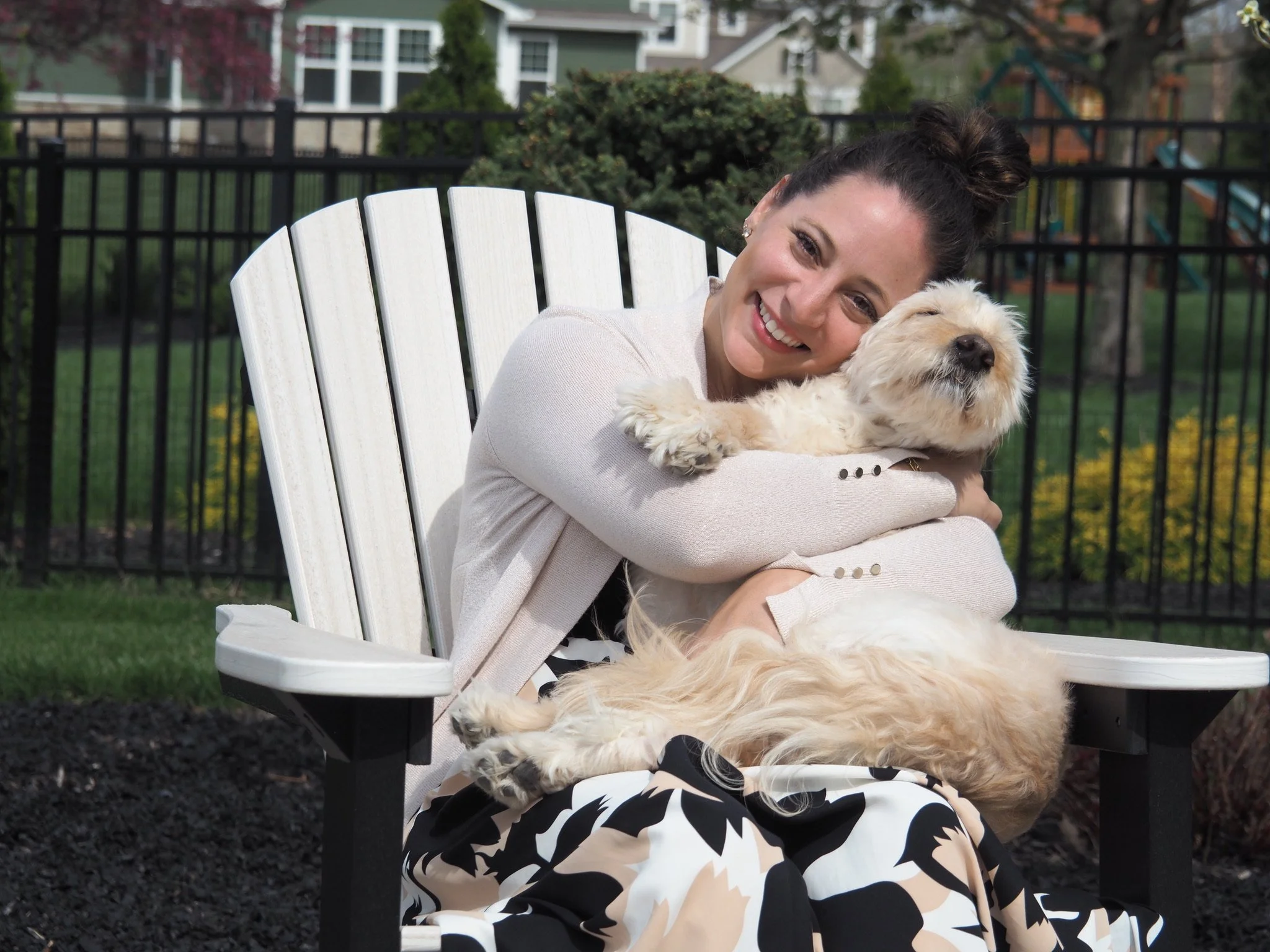 A woman with dark hair styled in a bun, wearing earrings and a neutral-colored blazer, sits on a white outdoor chair hugging a fluffy, light-colored dog with closed eyes against a background of a fenced yard and houses.