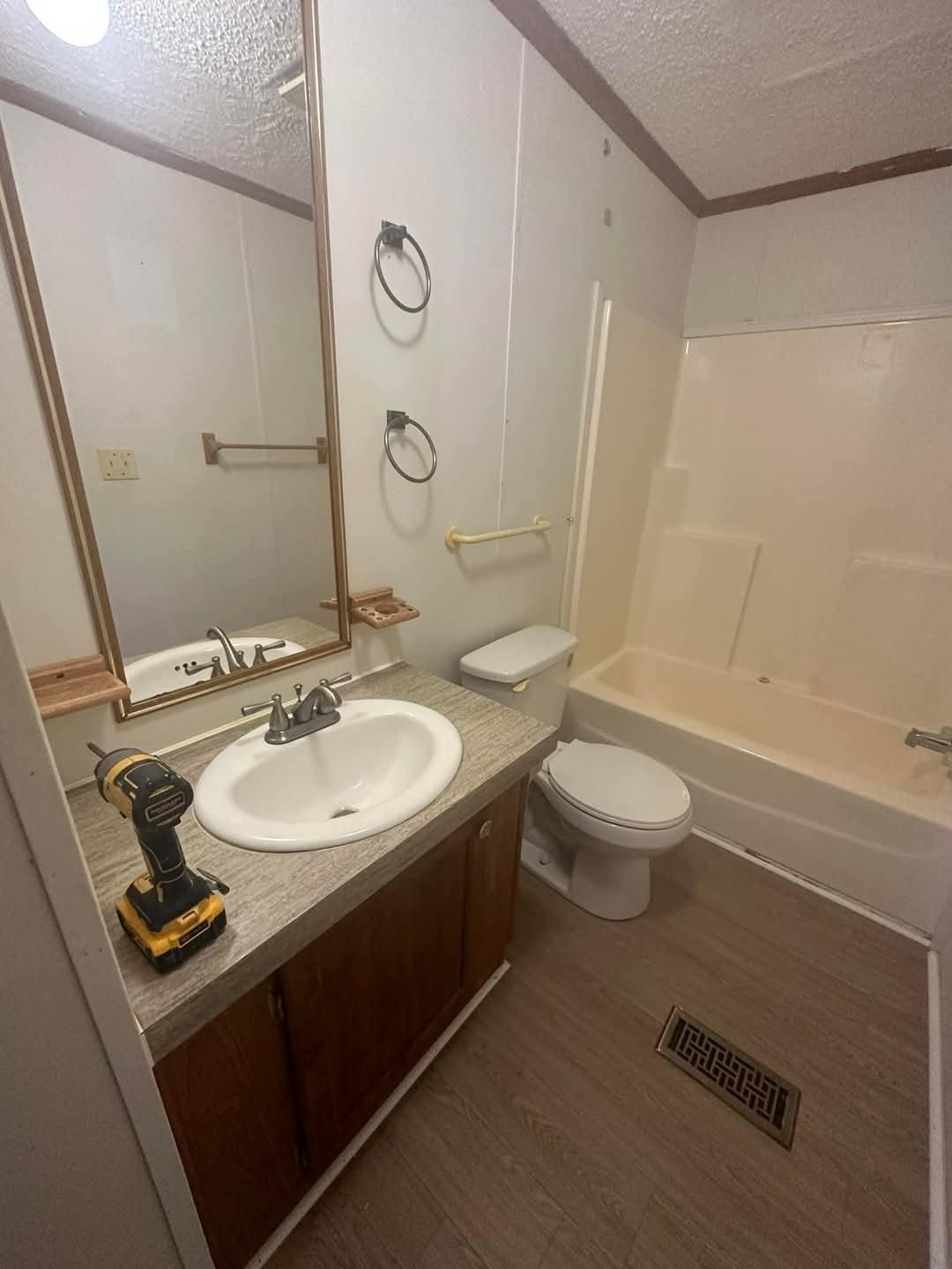 A small bathroom with a mirror, sink, and vanity cabinet. A cordless drill on the countertop, a toilet, a bathtub with a shower, and multiple towel rings and bars on the wall.