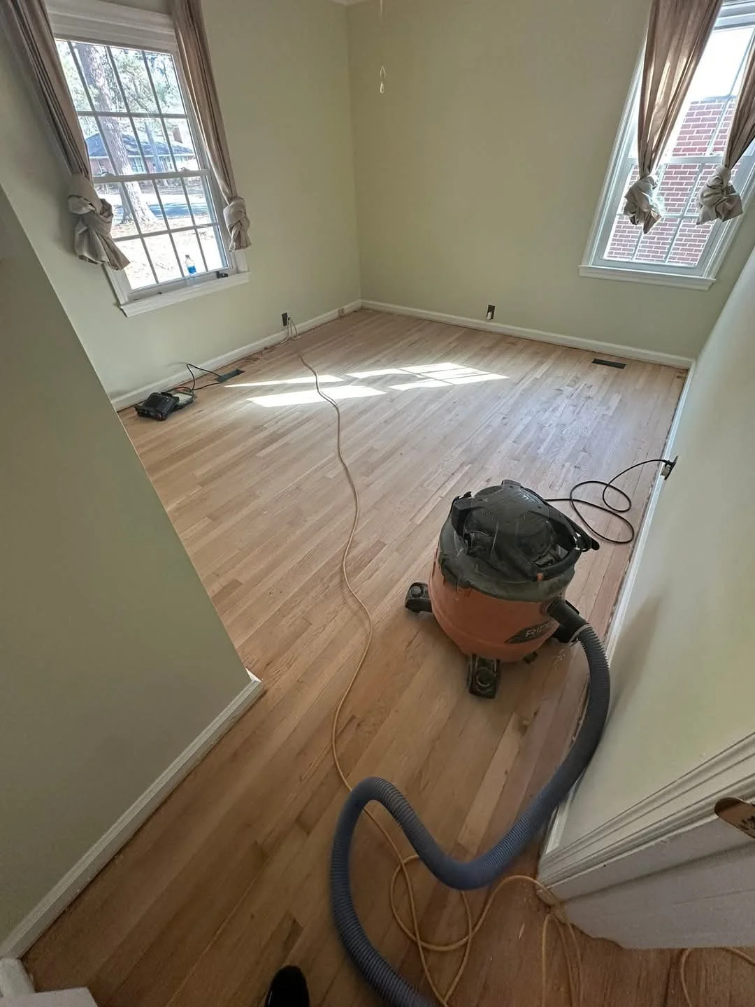 Empty room with light yellow walls and hardwood flooring. Two windows with beige curtains, letting in sunlight. A vacuum cleaner and an extension cord are on the floor, indicating cleaning or renovation in progress.