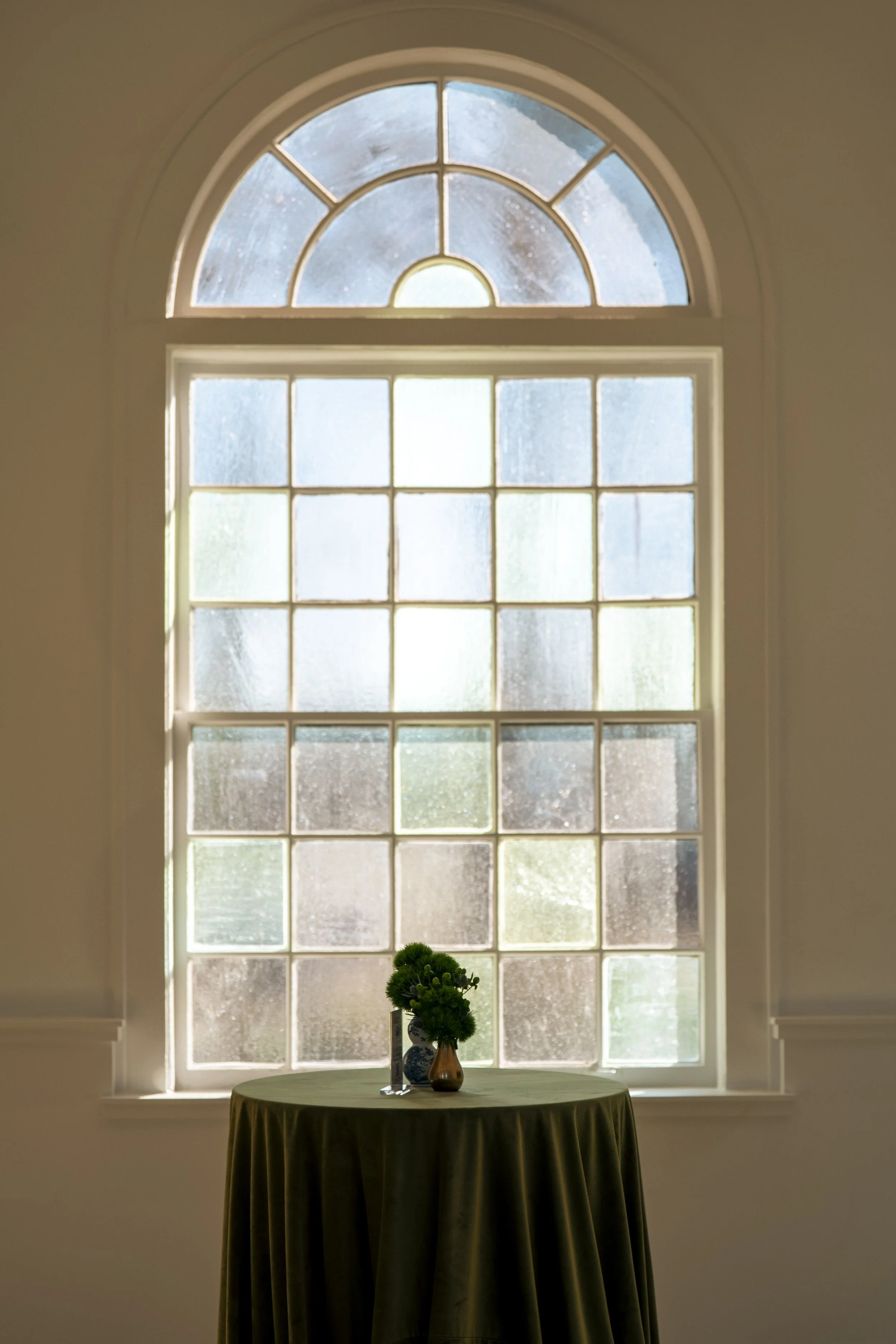 A round table covered with a dark green tablecloth, holding a small potted plant and decorative vases, in front of a large arched window with multiple panes and textured glass, allowing diffused sunlight to enter.