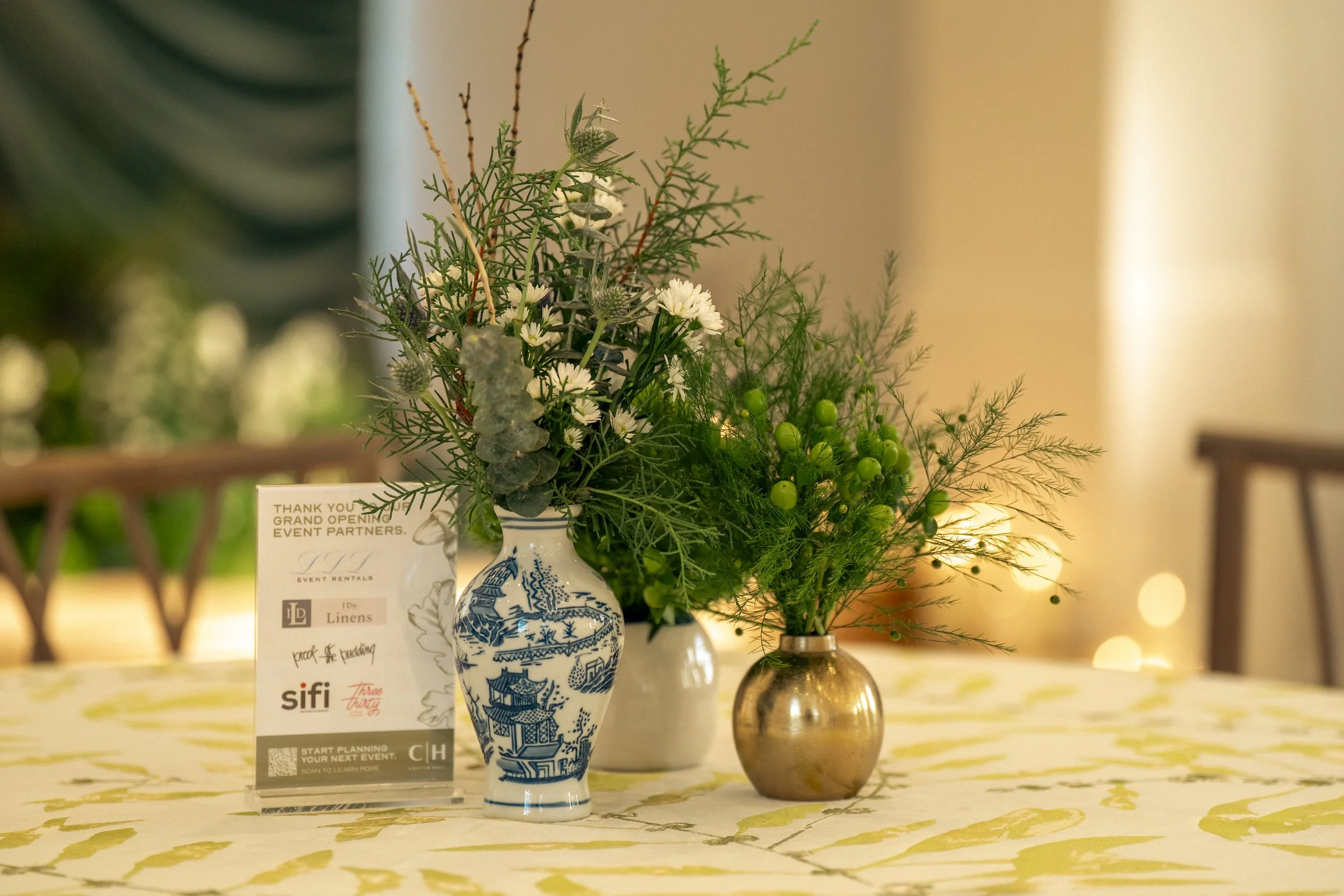 Elegant floral arrangement in blue and white porcelain vase, featuring white daisies, greenery, and twigs, on a table with event flyers and a gold-colored round vase in the background.