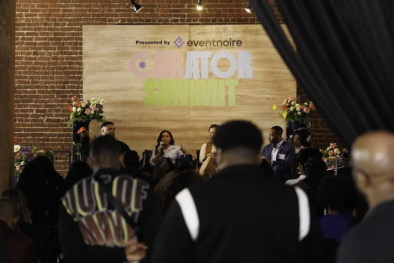 Panel discussion at the Curator Summit, with five speakers on stage and audience members listening in a brick-walled venue decorated with flowers, and a large banner with event branding in the background.