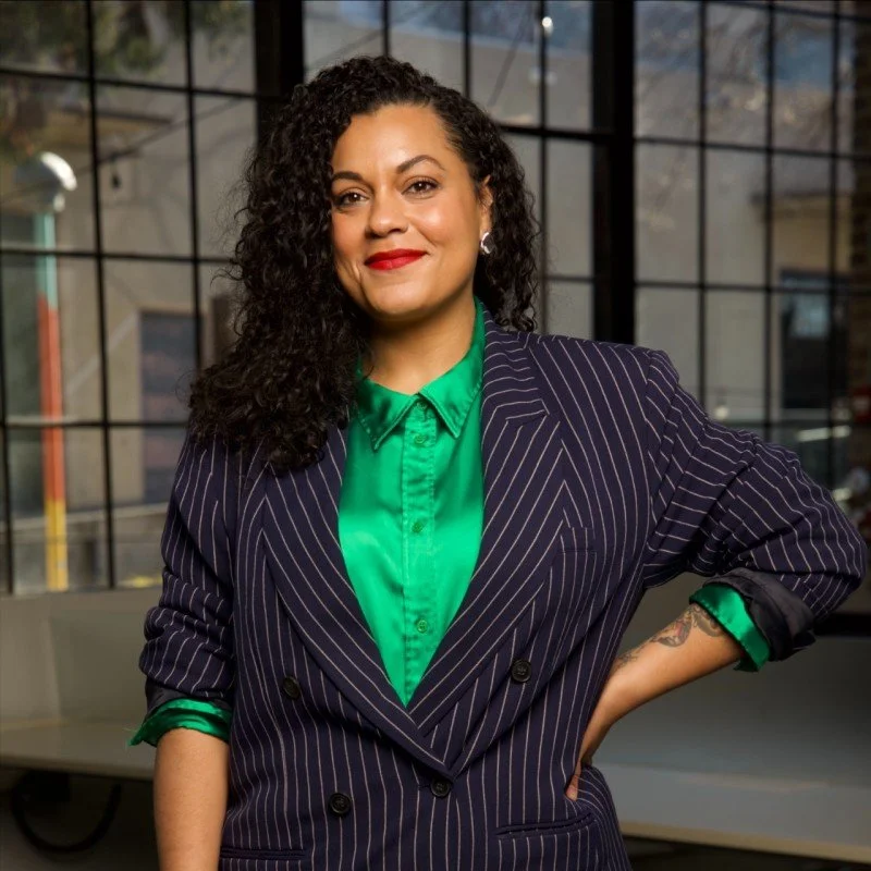 Confident woman with curly hair in a pinstripe blazer and green blouse standing indoors with large windows in the background.