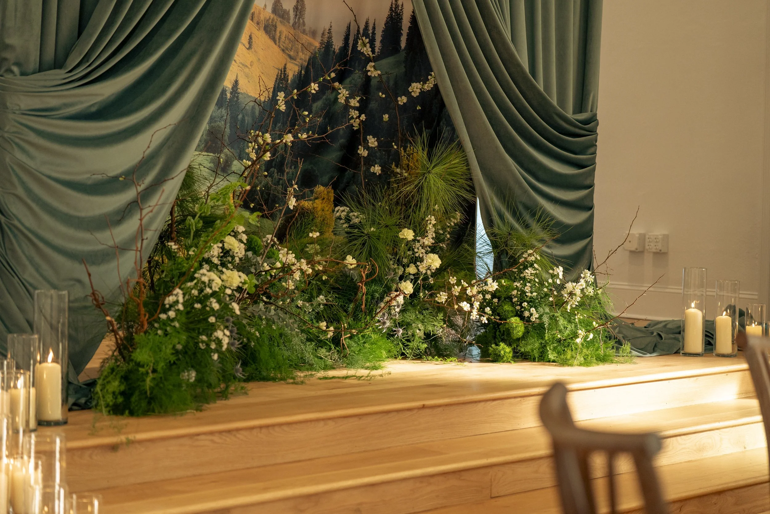 Decorative indoor setting with green curtains framing a backdrop of a mountain landscape mural, lush greenery, and white flowers, with lit candles on a wooden surface in the foreground.
