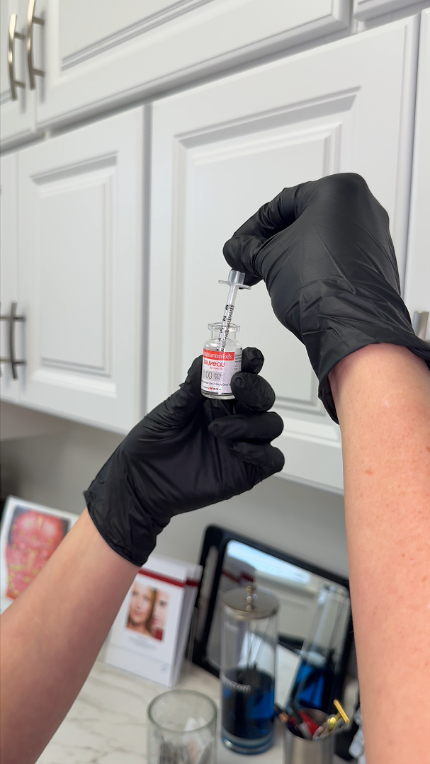 A person wearing black gloves using a syringe to draw a liquid vaccine from a small vial in a kitchen setting.