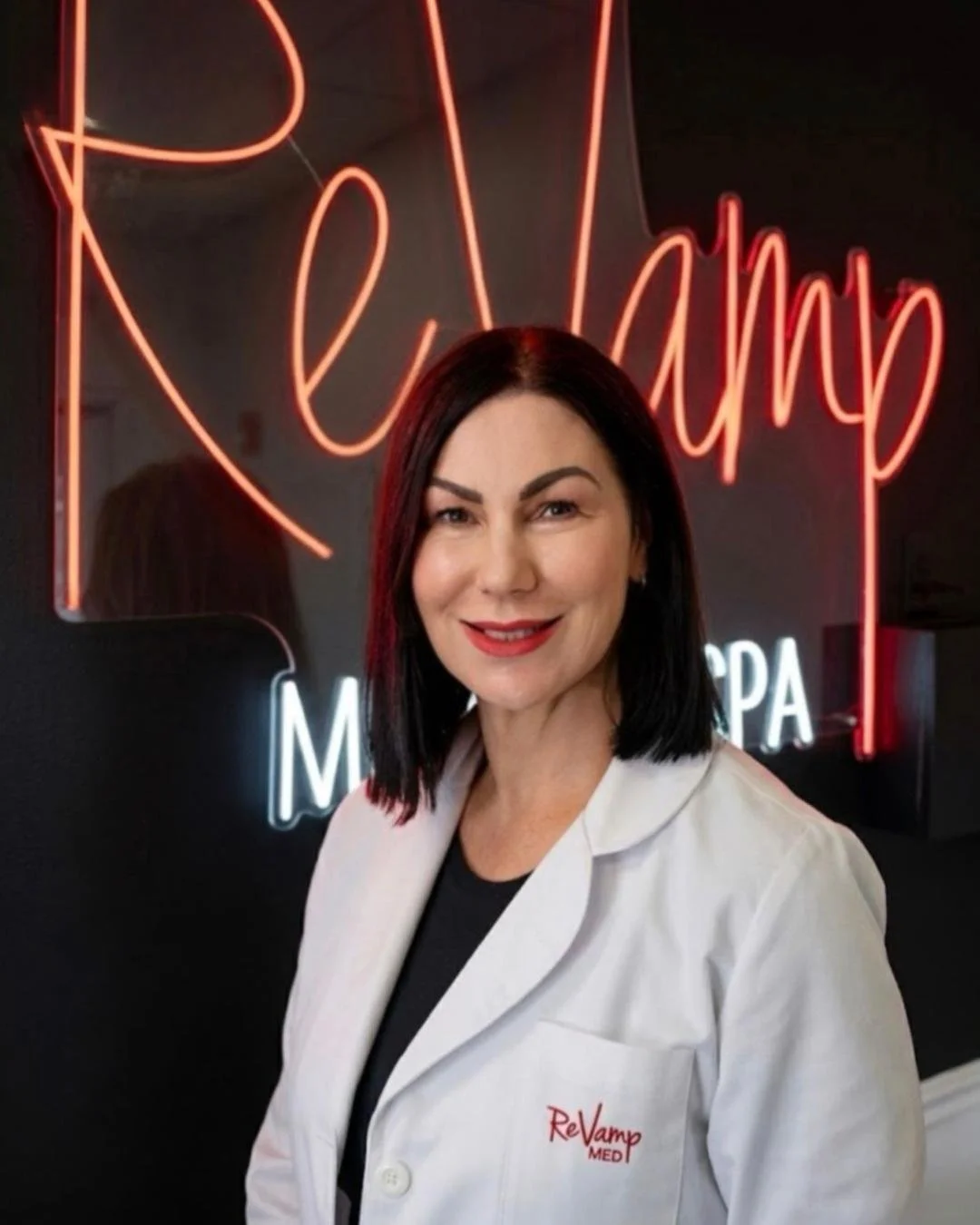 A woman with dark hair wearing a white medical coat with red embroidery standing in front of a neon sign that says "ReVamp" in red and white.