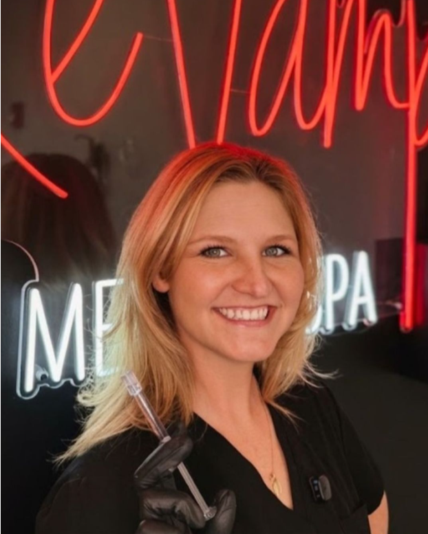 A smiling woman with blonde hair holding a pen, standing in front of a neon sign with red and white lights.