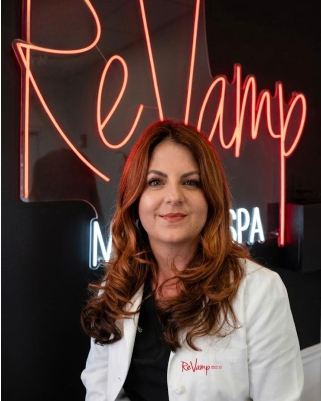 A woman with long red hair and a white lab coat stands in front of a neon sign that says "ReVamp."