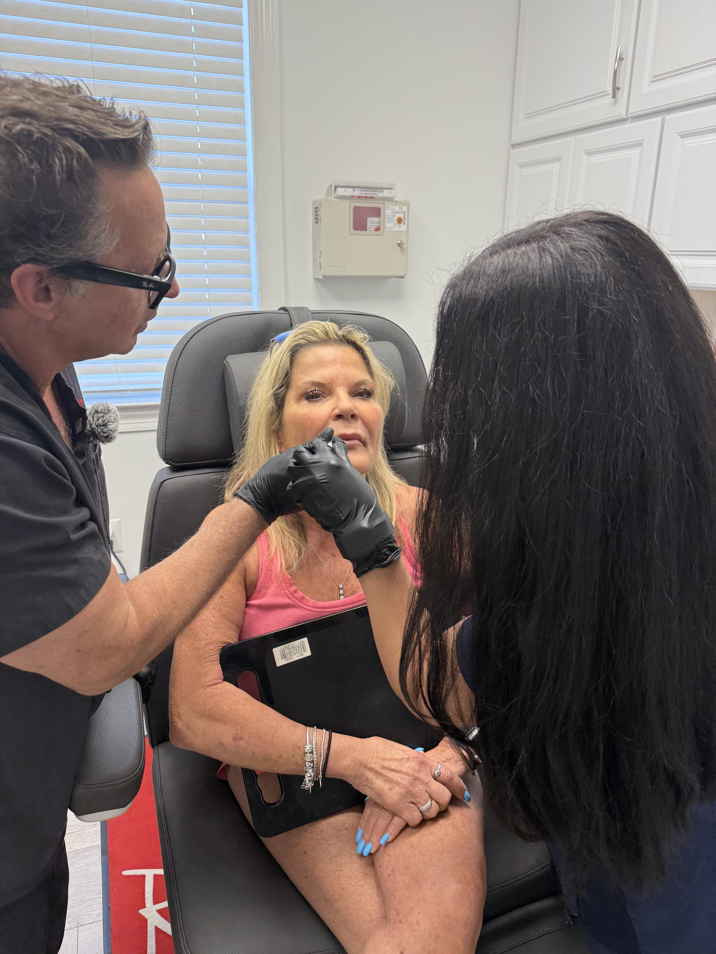 A woman receiving a cosmetic procedure on her face in a medical setting, with two healthcare professionals attending to her.