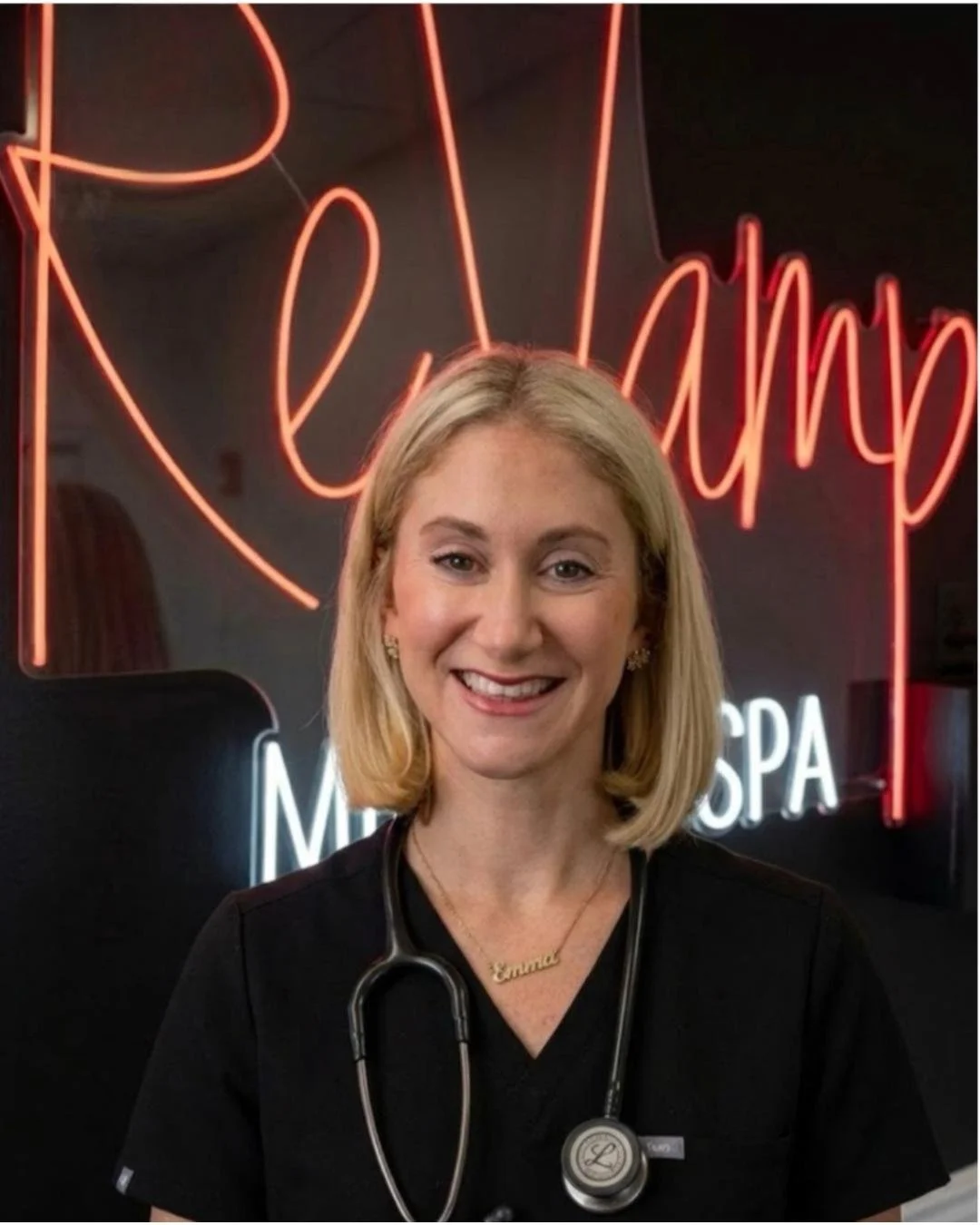 A smiling woman wearing a black medical uniform with a stethoscope around her neck and a gold necklace spelling 'Emma', standing in front of a neon sign that says 'Relax' and 'Med Spa'.