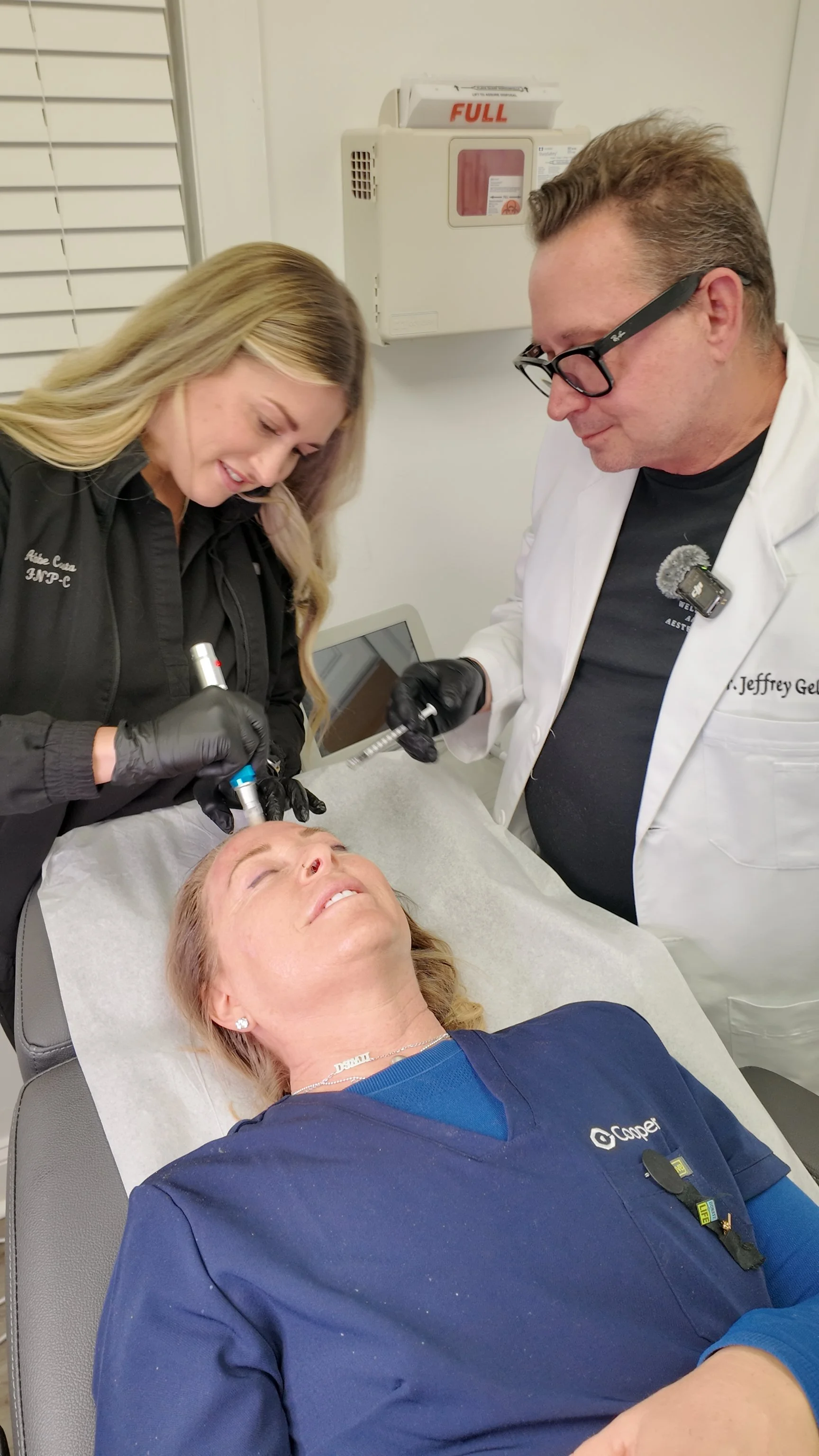 A woman lying on a medical chair with her eyes closed, receiving a facial treatment from a nurse, while a doctor stands nearby observing in a clinical setting.