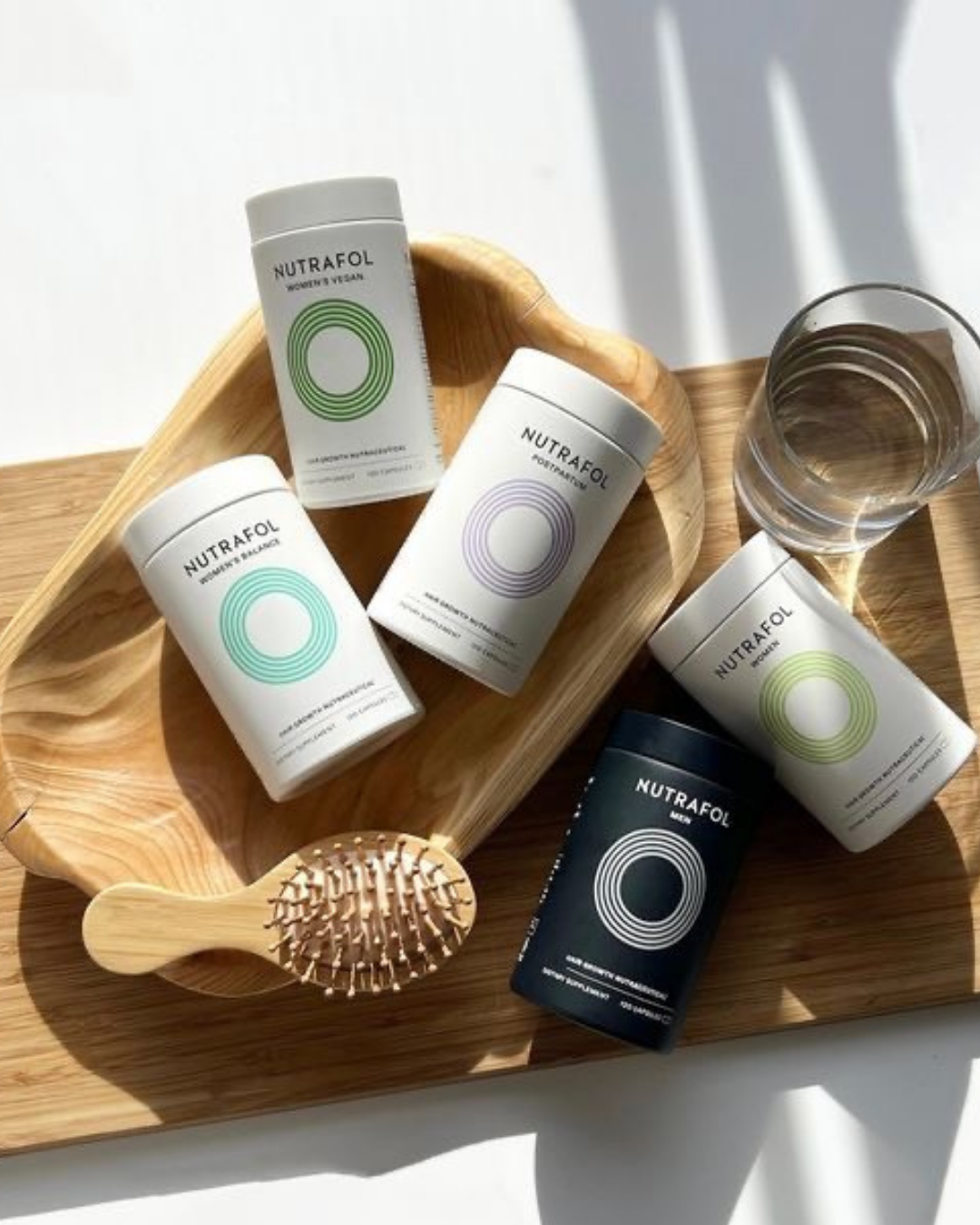 Collection of Nutrafol supplement bottles and a wooden hairbrush on a wooden tray with a glass of water, photographed in natural light.