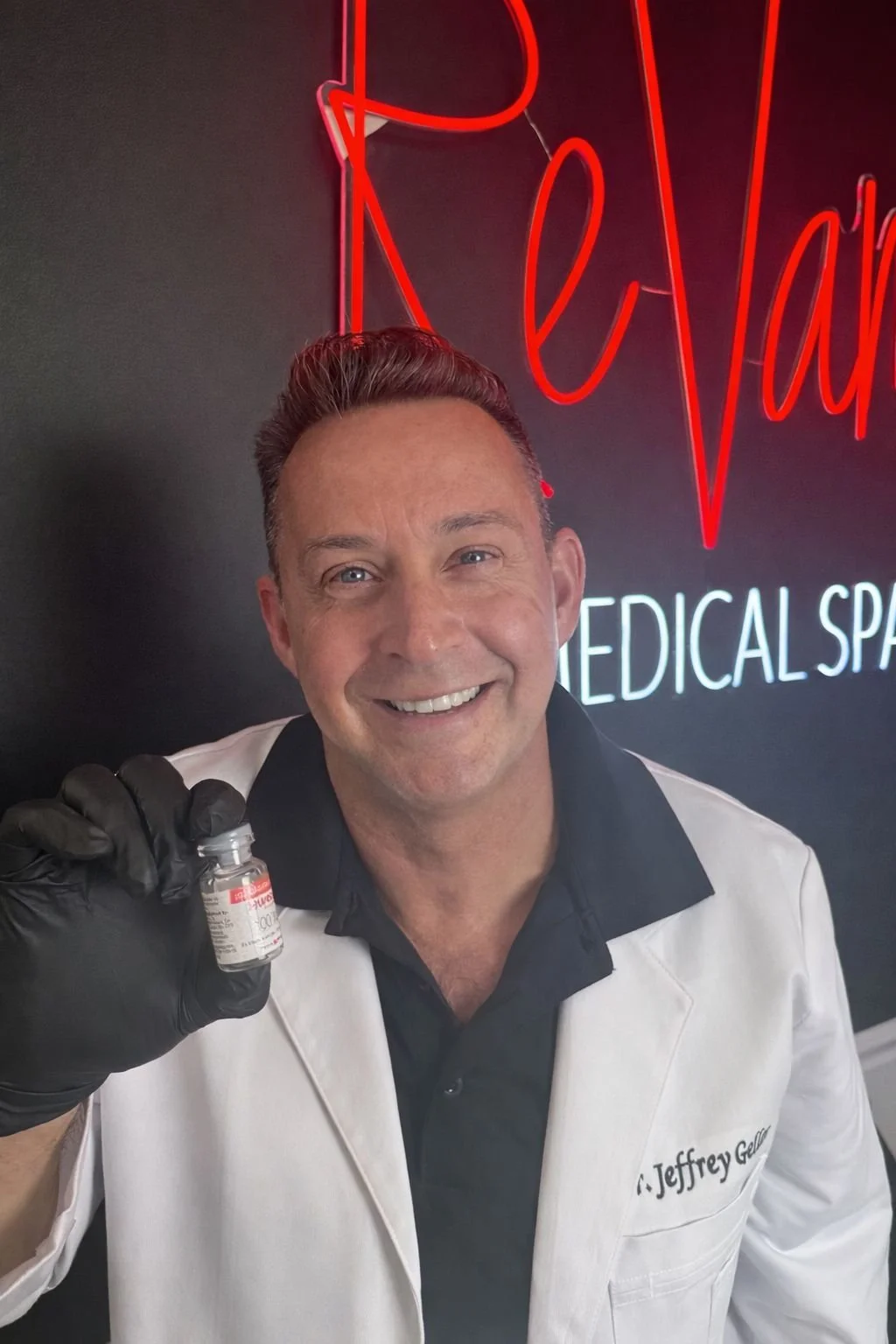 A smiling man in a white medical coat holding a small vial, standing in front of a neon sign that reads 'ReVamp Medical Spa'.