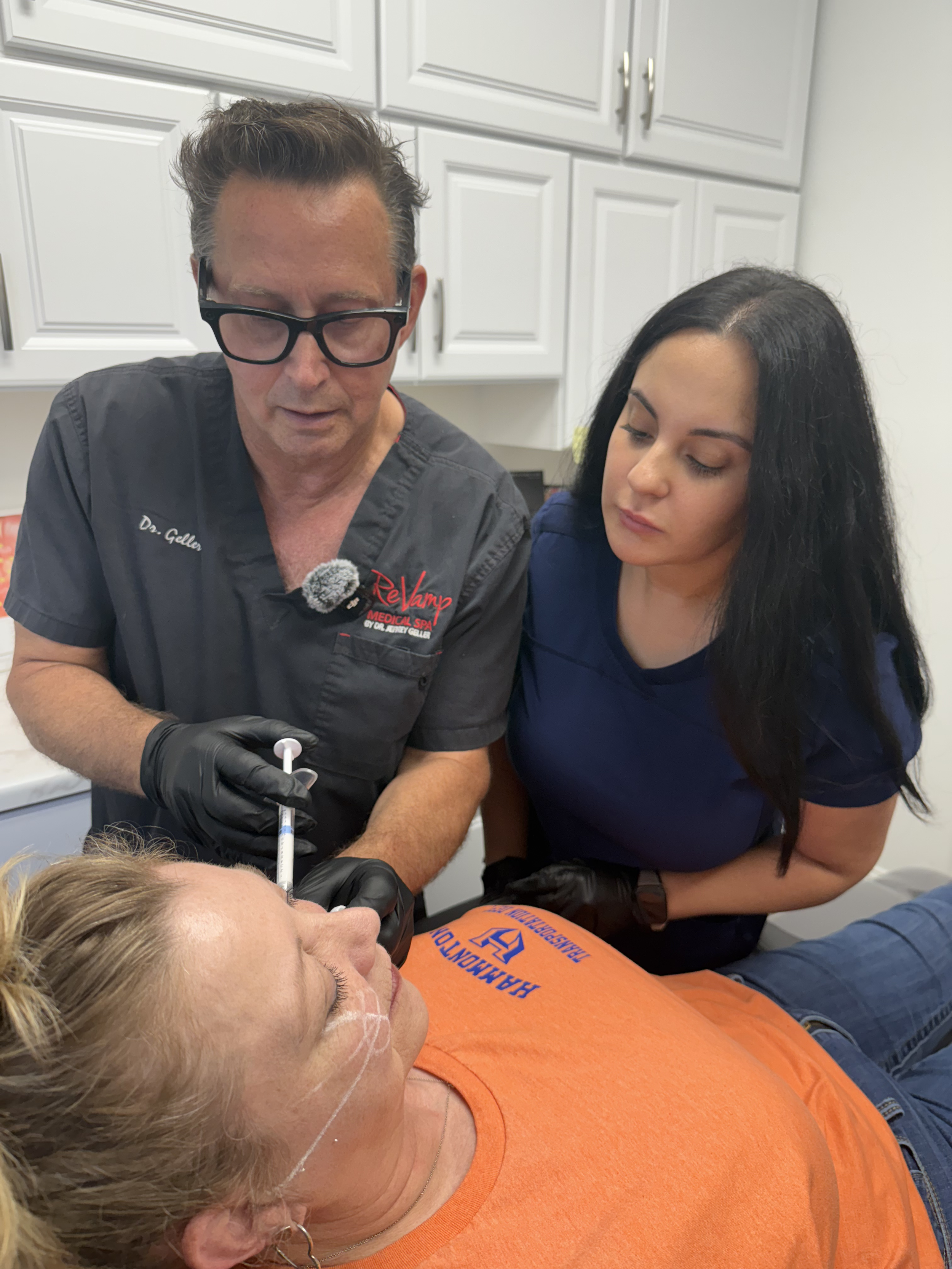 A medical professional prepares to give an injection to a woman lying on an examination table. The woman is wearing an orange shirt and has white lines drawn on her face, while another woman observes. The setting is a room with white cabinets.