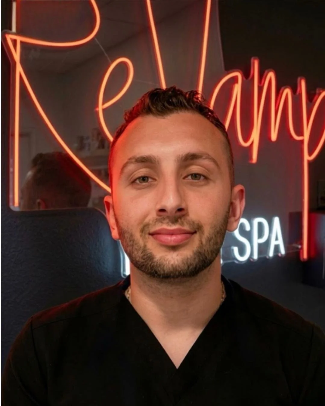 A young man with short dark hair and a beard, smiling slightly, is in front of a neon sign that says 'Relax' and 'SPA' in a spa or salon setting.