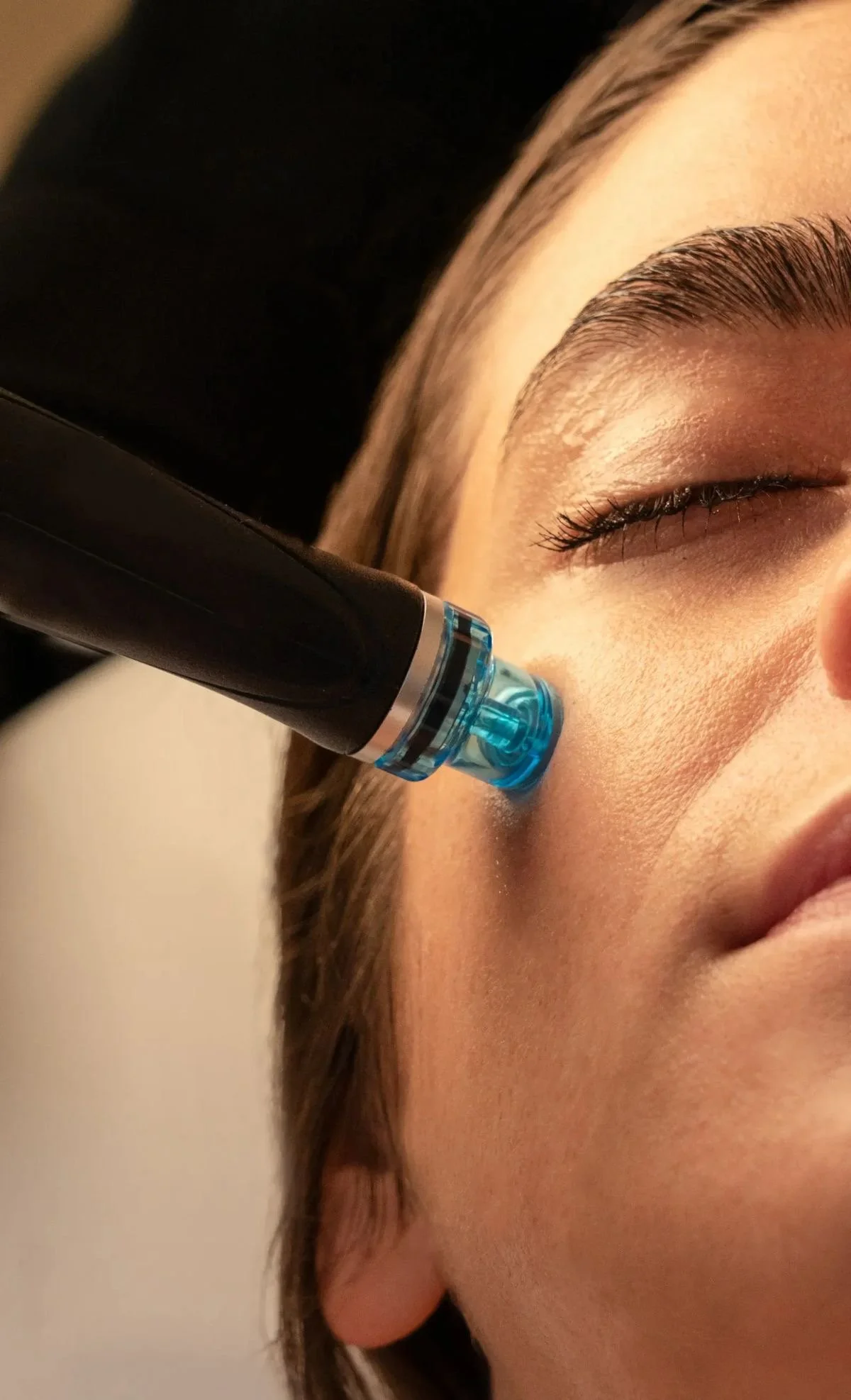 Close-up of a woman receiving a facial treatment with a microdermabrasion device on her cheek, eyes closed, with smooth skin.