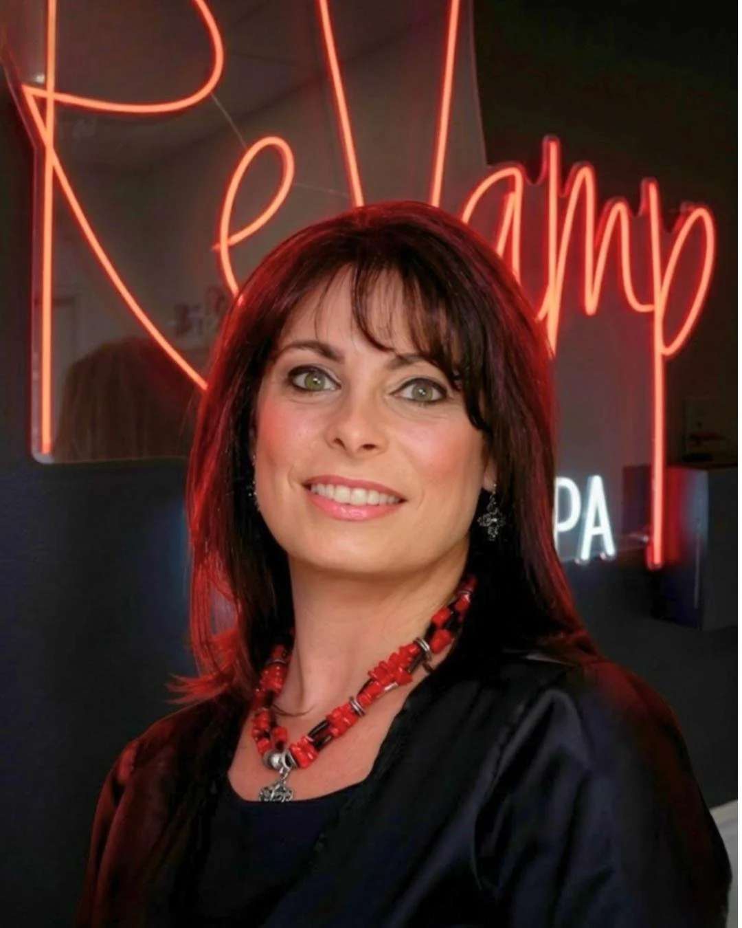 Portrait of a woman with dark red hair, wearing a black top and red necklace, smiling in front of a neon sign that reads 'relax' with red and white lighting.
