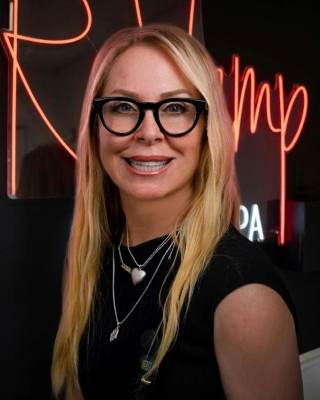 A woman with long blonde hair, wearing glasses, multiple necklaces, and smiling, standing in front of a black wall with a red neon sign in the background.