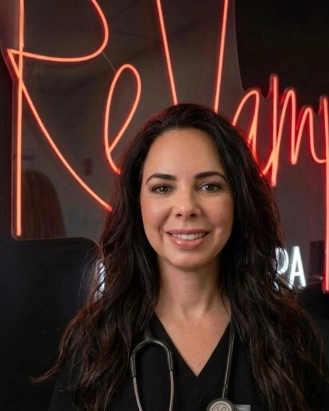 A smiling woman with long dark wavy hair and a stethoscope around her neck, standing in front of a neon sign that says 'Read' in cursive.
