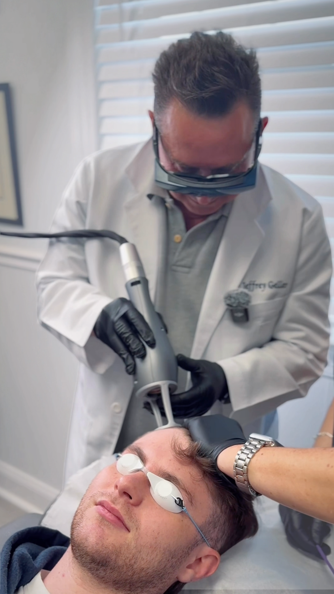 A medical professional performs a procedure on a male patient lying down, with eye protection on the patient and wearing black gloves, in a clinical setting.