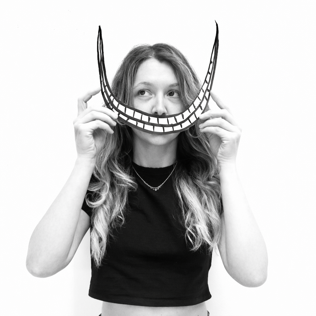 Young woman with long wavy hair holding a cutout of a smiling mouth mask in front of her face, against a plain background.