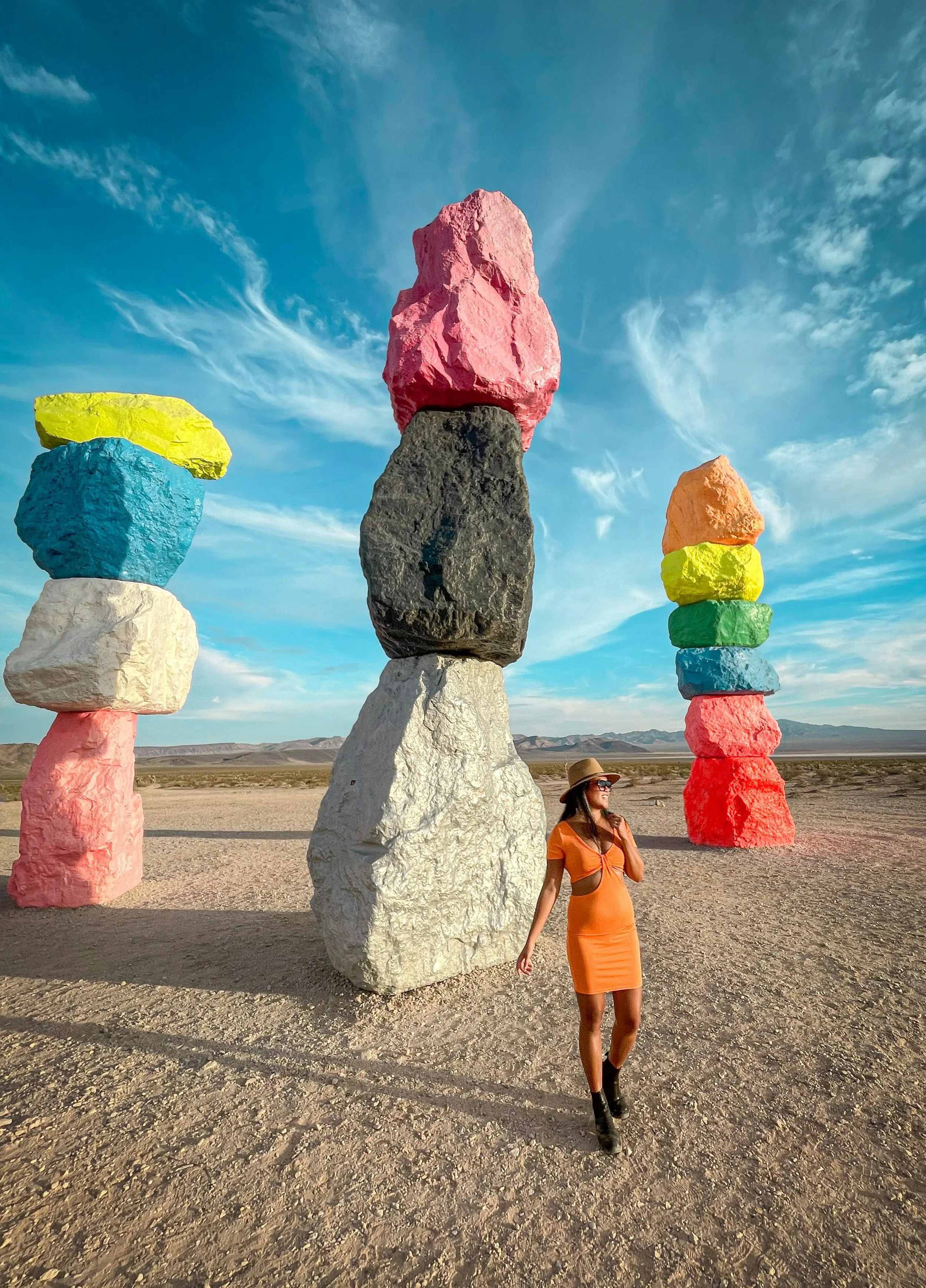 A woman in an orange dress and hat walking near colorful stacked rocks in a desert landscape under a blue sky.