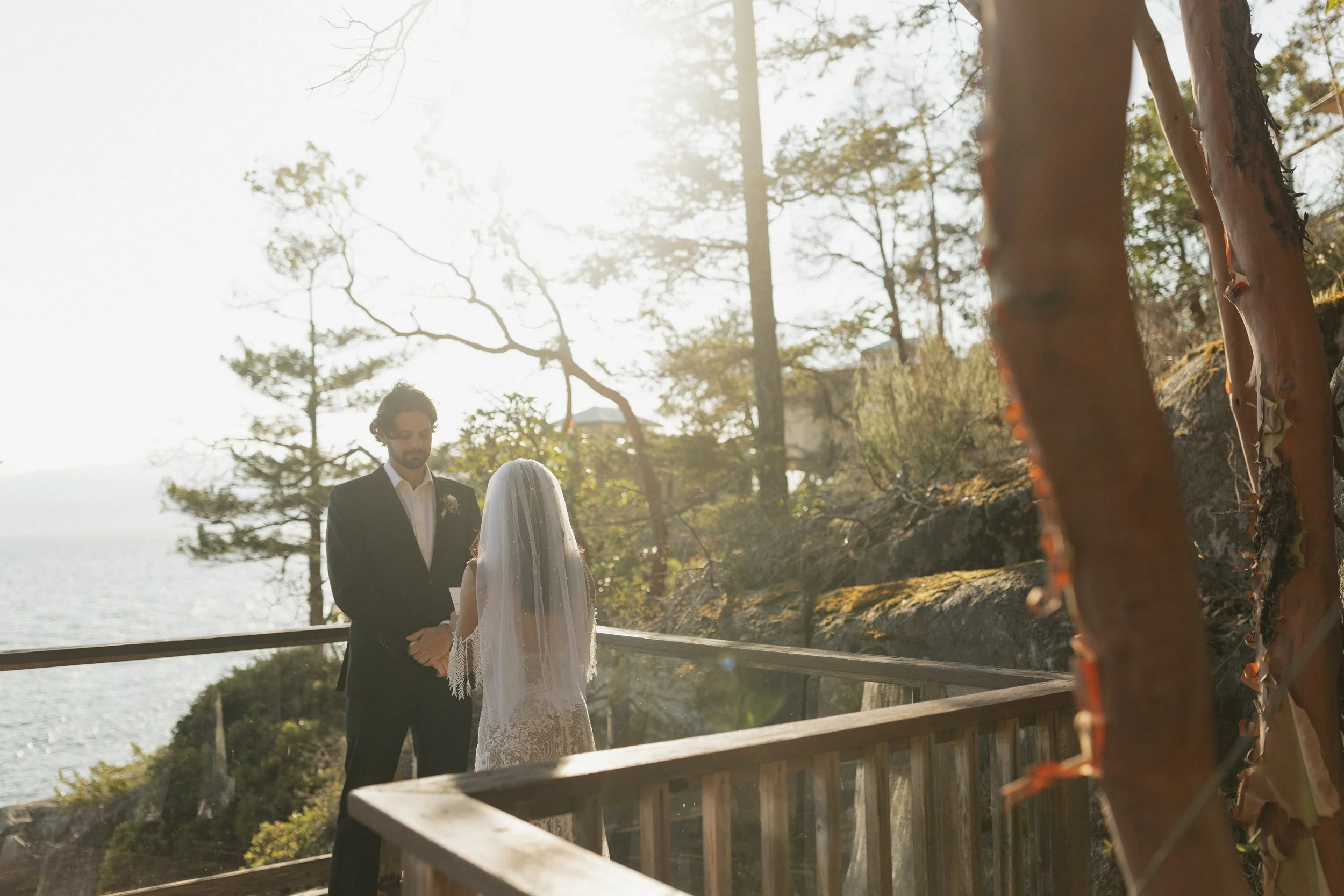 A couple is standing outdoors during a ceremony, with a woman officiating and holding a certificate or book. The woman on the left has long hair with tattoos on her arms and is holding a bouquet of flowers. The man on the right is wearing a white shirt with rolled-up sleeves and is looking at the woman officiating. The background features trees and bright sunlight.