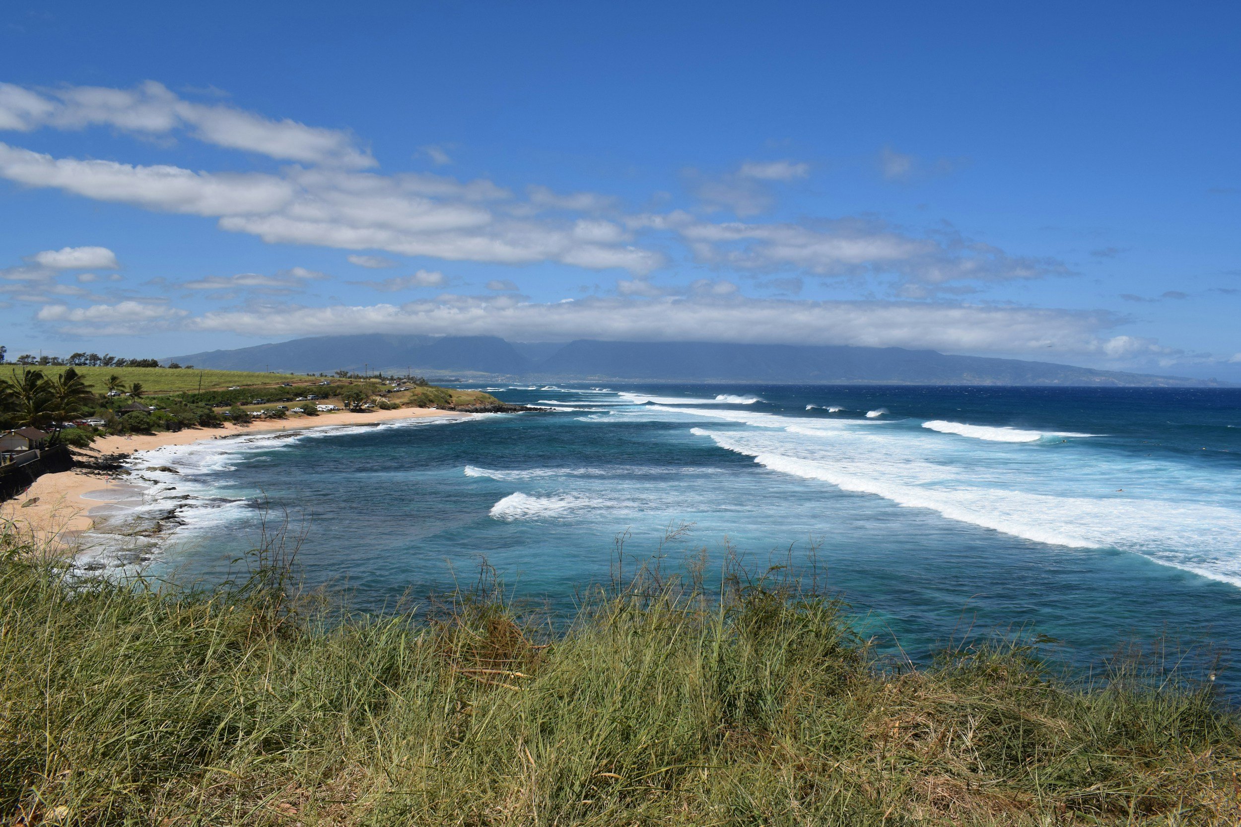 View of a tropical beach with grassy foreground, sandy shoreline, blue ocean waves, distant mountains, and a partly cloudy sky.